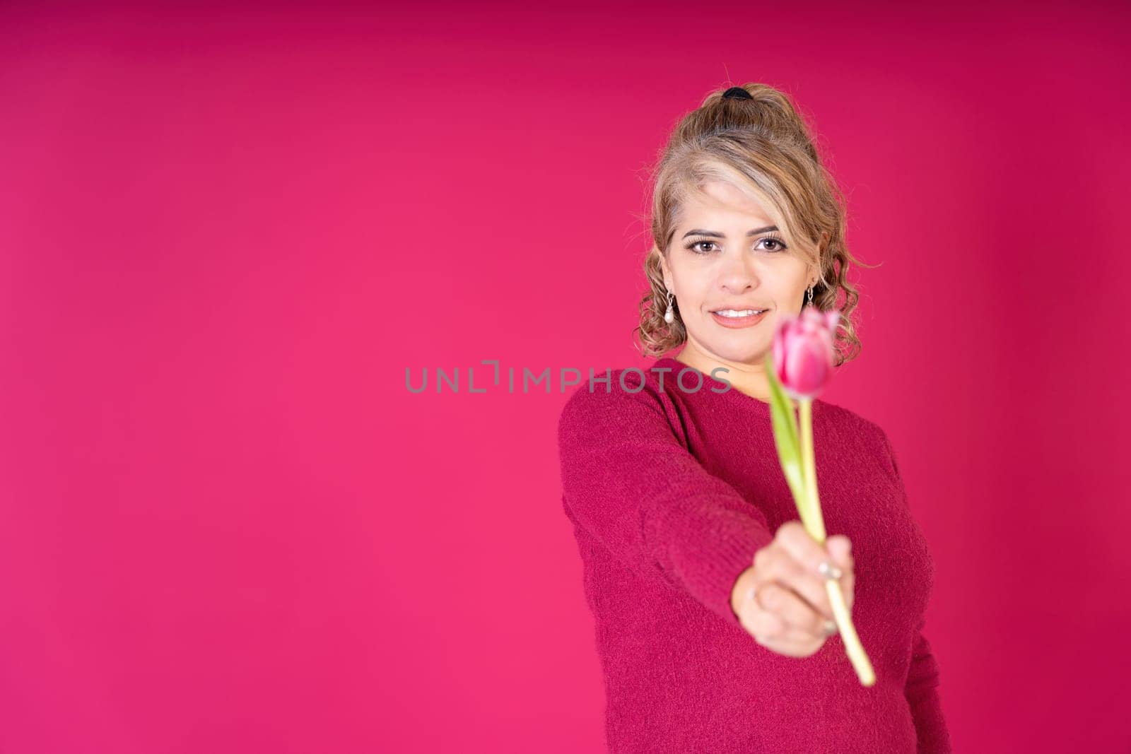 Young pretty woman smiling looking at camera offering a tulip flower, wearing a red t-shirt on a red background with copy space. Gifts concept.