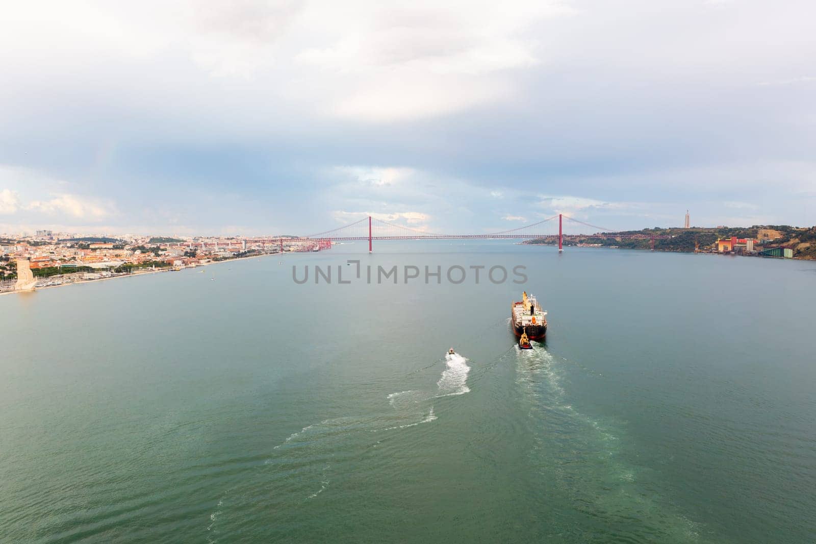 Aerial drone shot of cargo ship and longboat moving in calm sea along mountains under cloudy sky, import export around the world