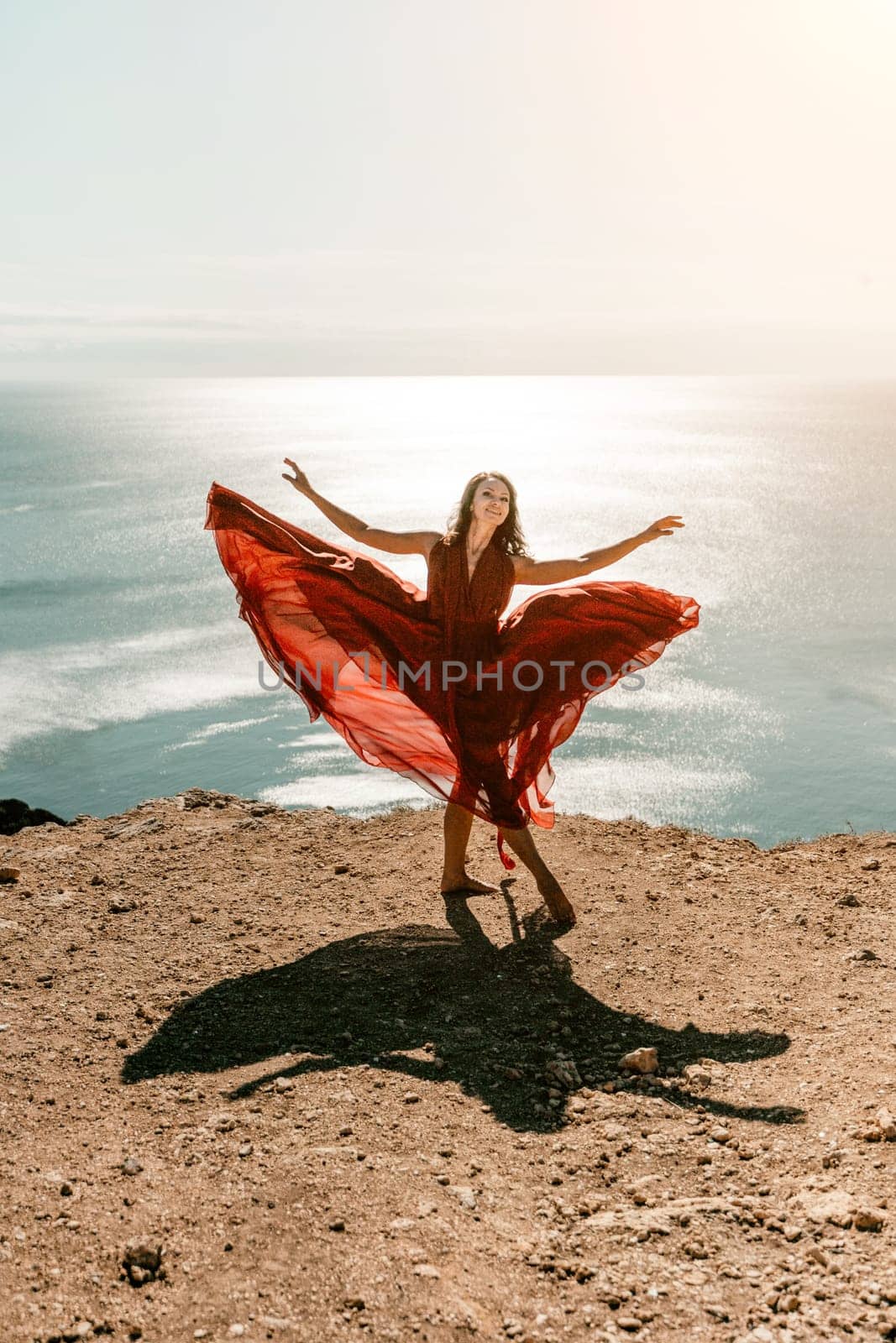 Woman red dress sea. Female dancer in a long red dress posing on a beach with rocks on sunny day. Girl on the nature on blue sky background