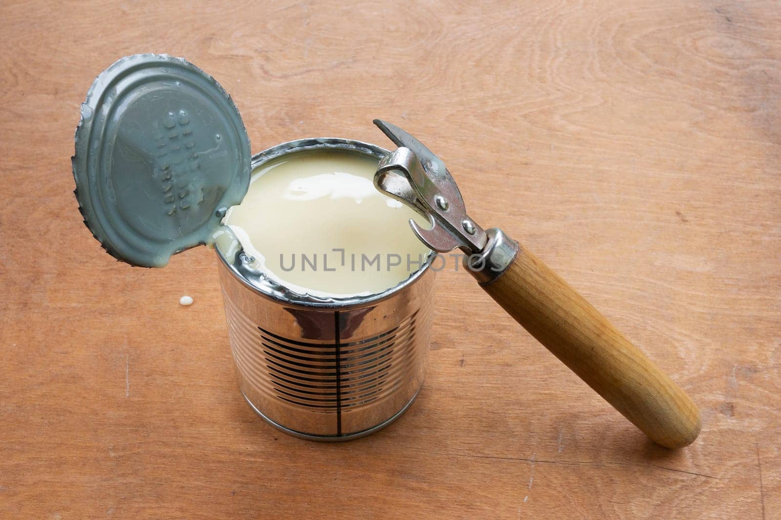 Opened metal can of condensed milk and metal can opener on brown wooden background. Camping food concept.