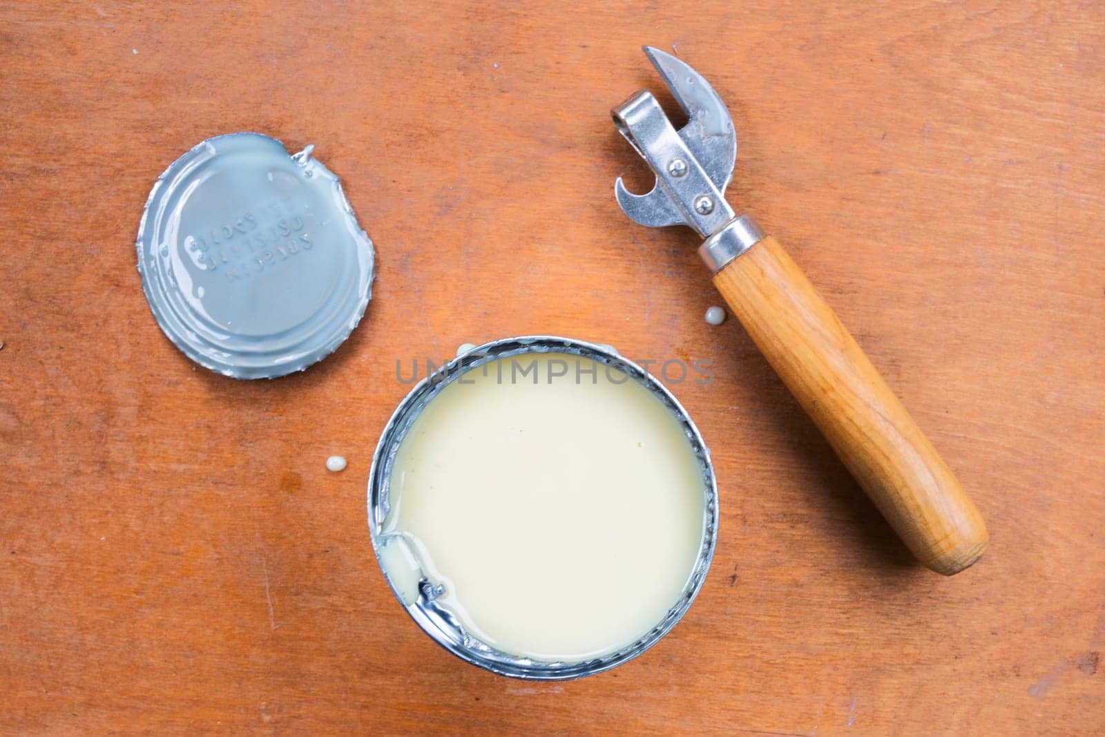 Open metal can with condensed milk, cut off lid, can opener on brown background, top view.