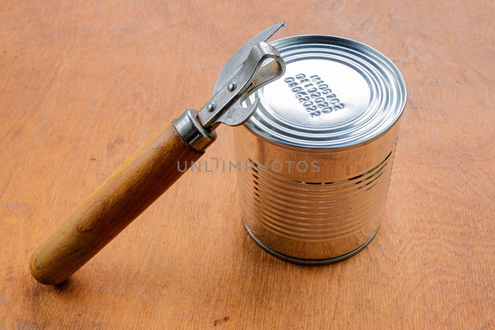 Unopened metal can and can opener on brown wooden background.