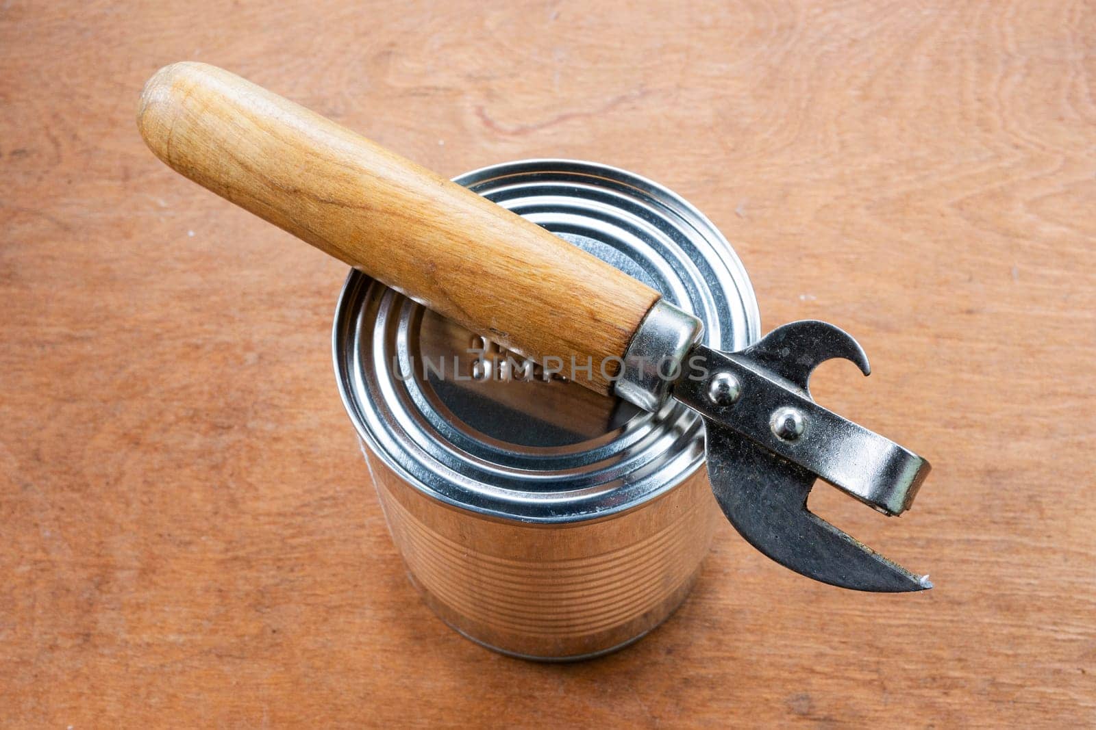 Camping food concept. Unopened metal can and can opener on brown wooden background.