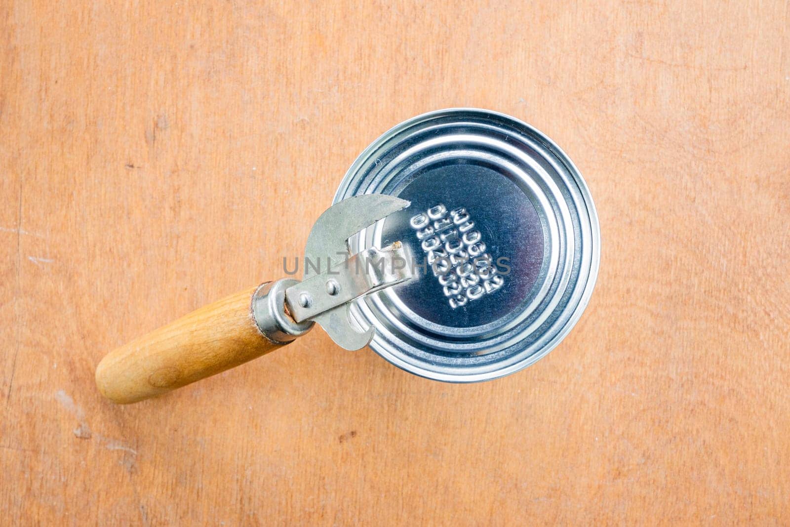 Unopened metal can and can opener on wooden brown background, top view.