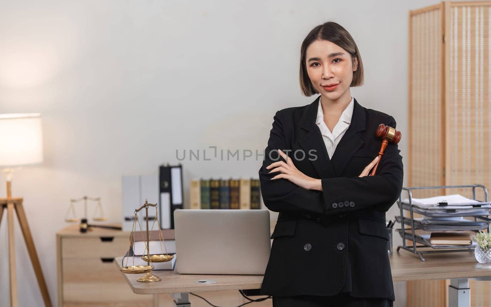 Confident young lawyer standing in modern office, holding gavel, with legal books and scales of justice in the background.