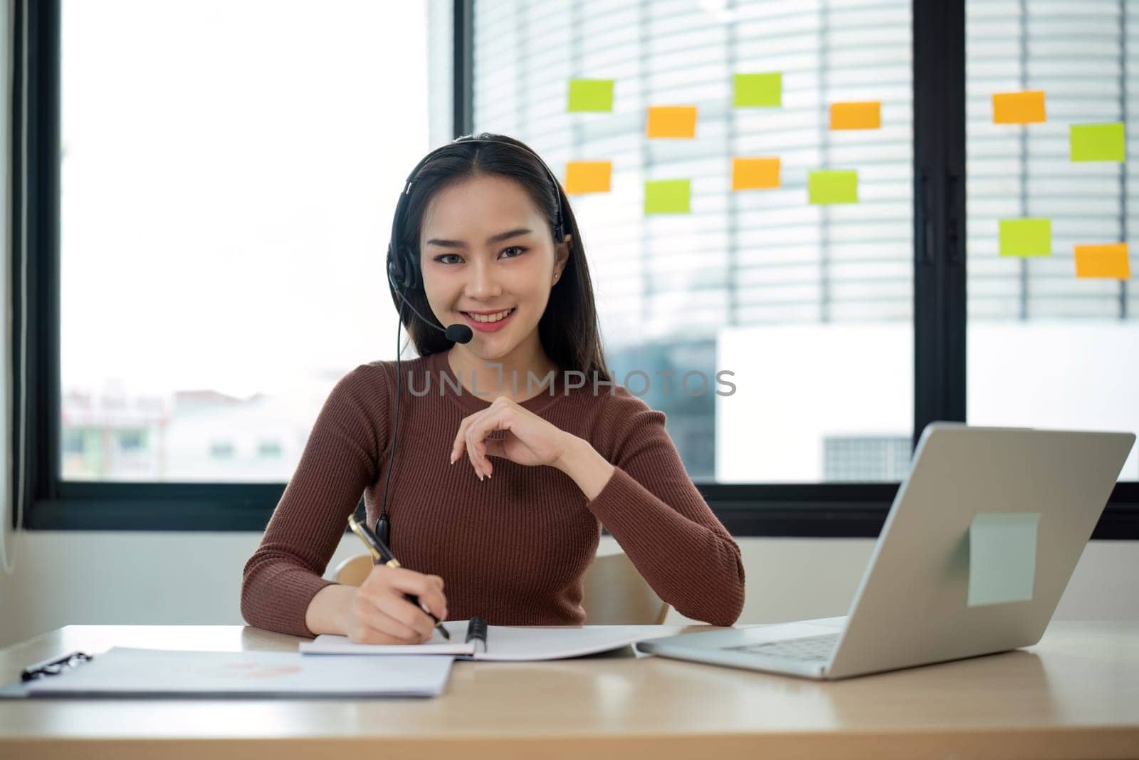 A woman wearing a headset is sitting at a desk with a laptop and a notebook by wichayada