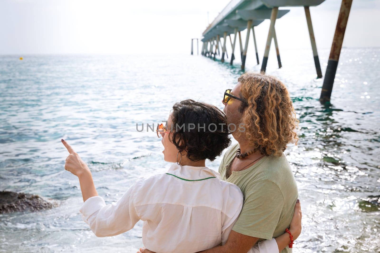 Smiling couple in sunglasses enjoying vacation hugging each other pointing and looking at the sea.
