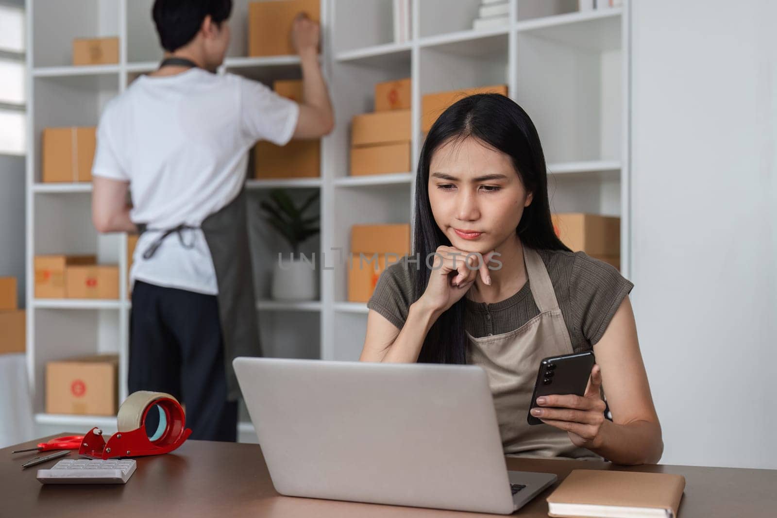 A couple feeling stressed while managing their online sales business from home, using a laptop and smartphone, surrounded by shipping boxes.