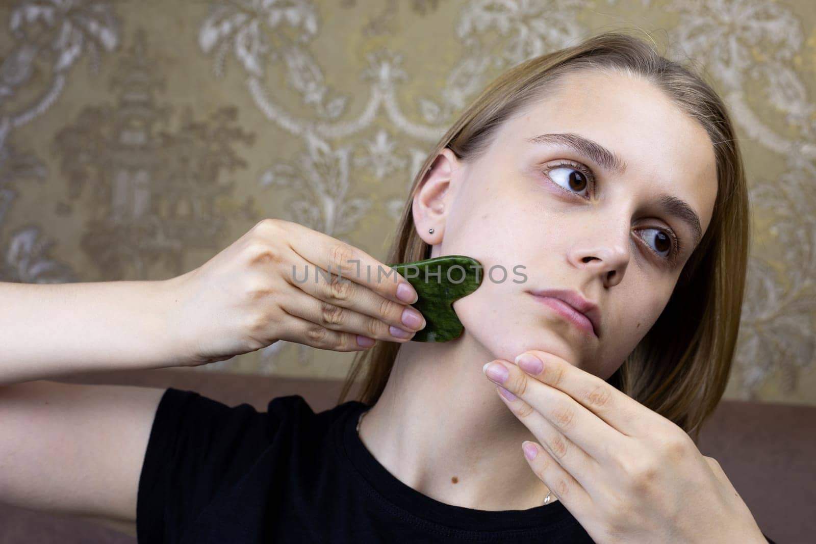 European girl stretches wrinkles on her facial skin using gua sha scraper made of natural jade colored stone, procedure at home for cleaning and caring for skin of young girl face