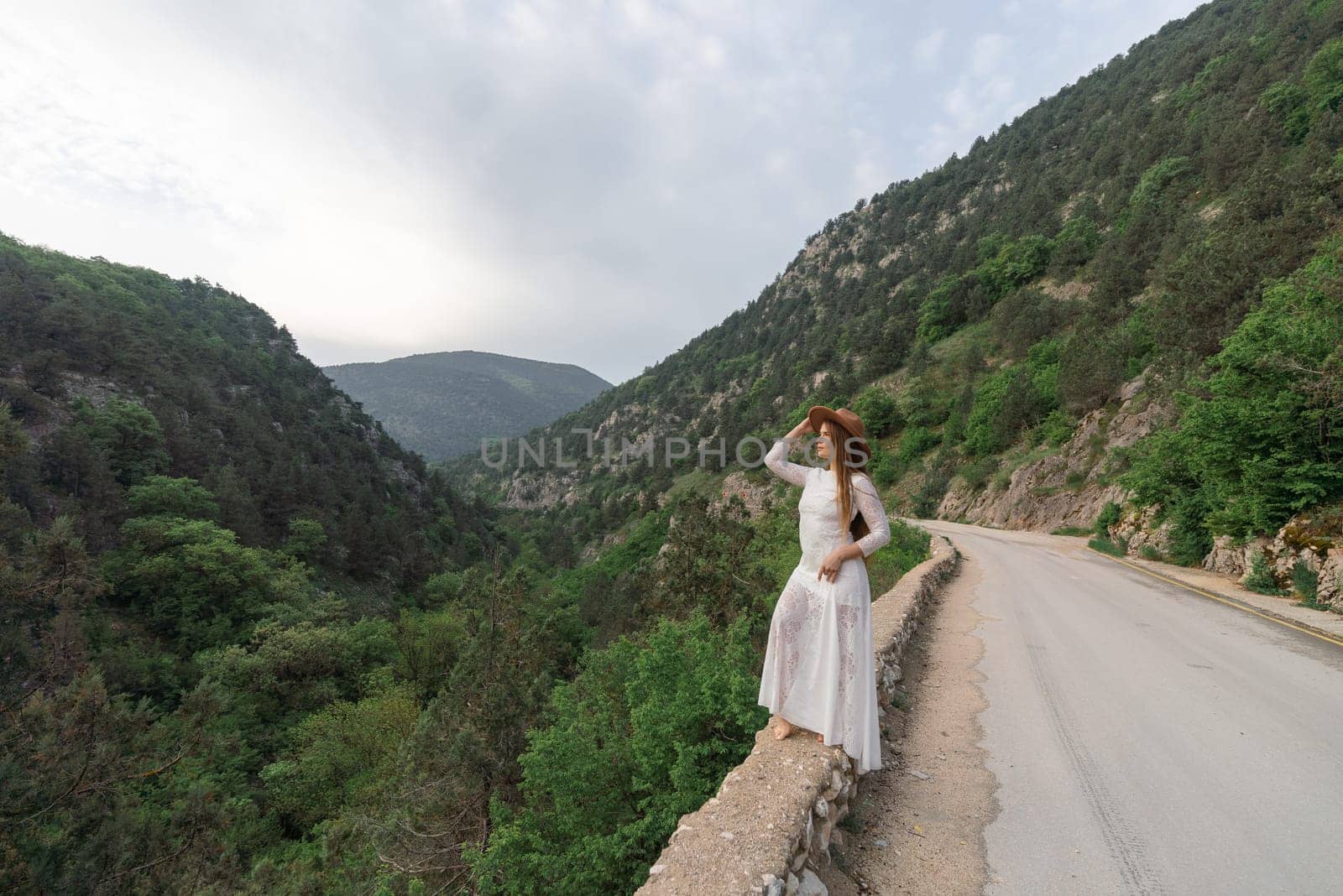 A woman in a white dress stands on a road overlooking a mountain range. The scene is serene and peaceful, with the woman's hat adding a touch of whimsy to the landscape
