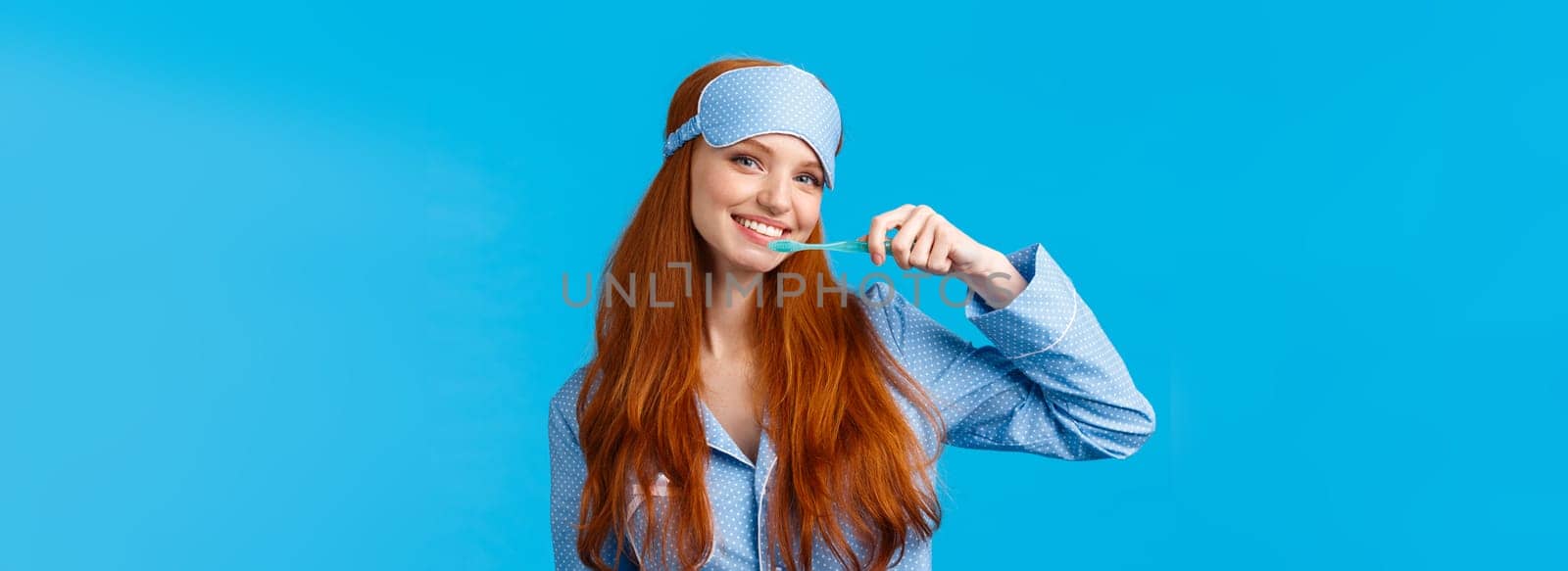 Cheerful cute redhead woman in nightwear, pyjama and sleep mask, brushing teeth holding toothbrush and smiling cheerful, wake up and taking care hygiene, standing blue background.