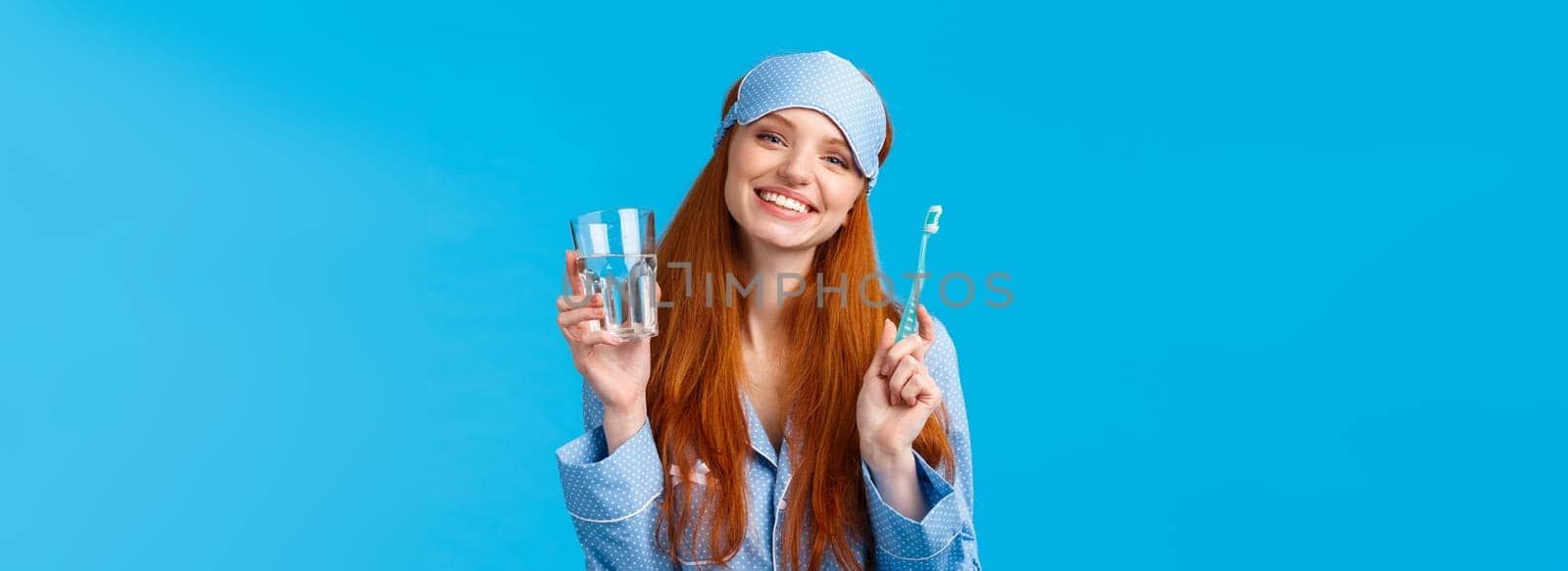 Happy smiling and carefree redhead enthusiastic girl in sleep mask, pyjama, holding glass water and toothbrush, taking care mouth hygiene, dental daily routine, blue background.
