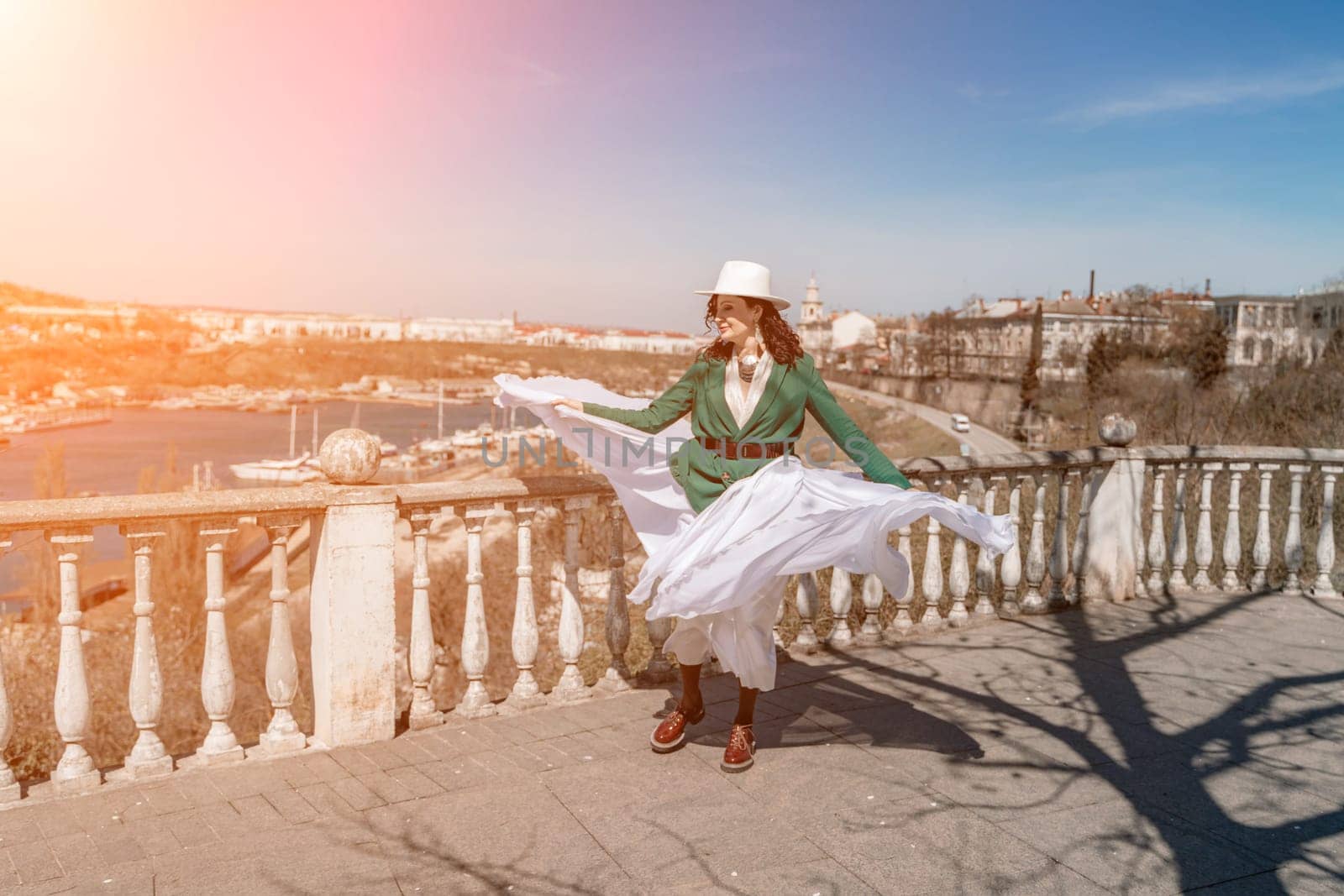 Woman walks around the city, lifestyle. Happy woman in a green jacket, white skirt and hat is sitting on a white fence with balusters overlooking the sea bay and the city. by Matiunina