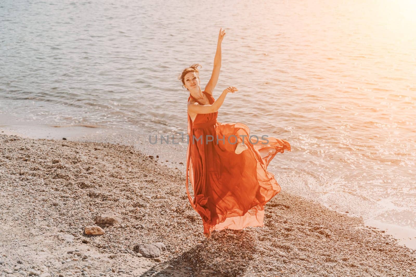Woman red dress sea. Female dancer in a long red dress posing on a beach with rocks on sunny day. Girl on the nature on blue sky background