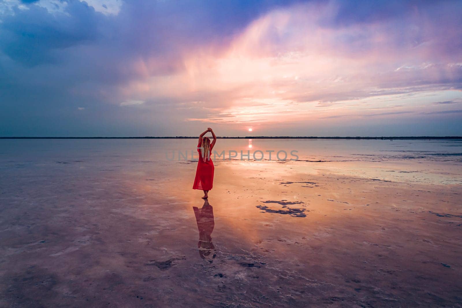 A woman in a red dress stands on the shallow waters of Burlinskoe Lake as the sun sets over the horizon, creating a beautiful reflection on the water.