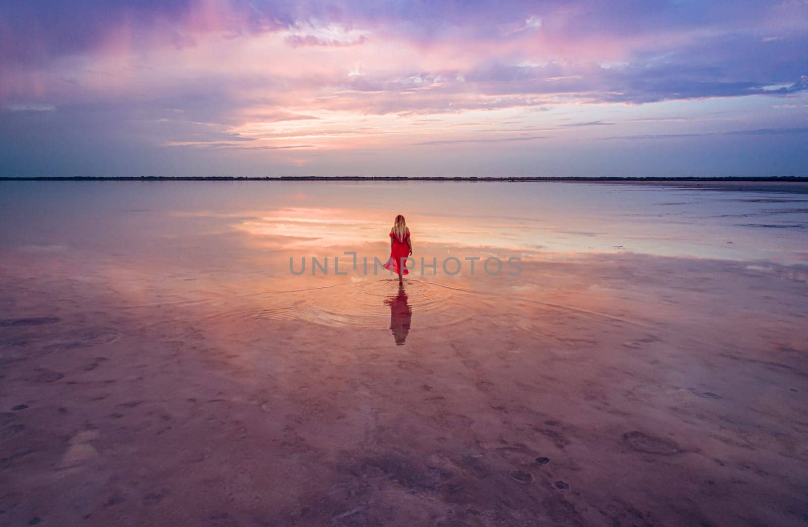 Aerial of a young woman in red dress walking in the water of a unique pink salt lake. Sunset at lake Bursol with beautiful reflections on calm water surface. Stunning scenery.