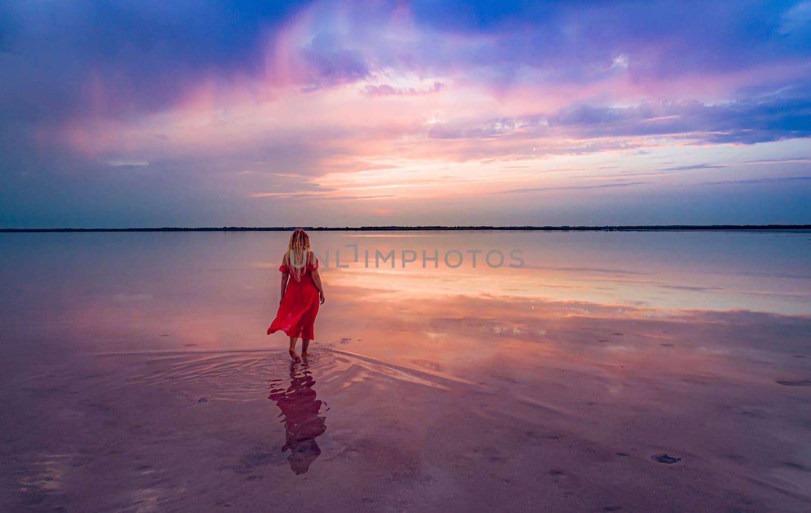 Aerial of a young woman in red dress walking in the water of a unique pink salt lake. Sunset at lake Bursol with beautiful reflections on calm water surface. Stunning scenery.