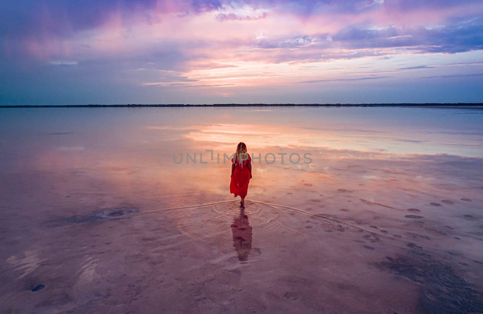 Aerial of a young woman in red dress walking in the water of a unique pink salt lake. Sunset at lake Bursol with beautiful reflections on calm water surface. Stunning scenery.