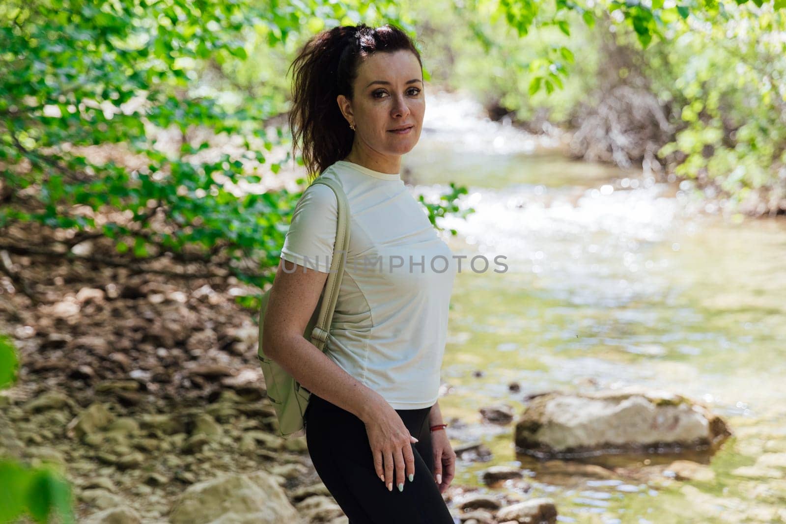 woman with a backpack on a hike stands by the river in a forest of green trees