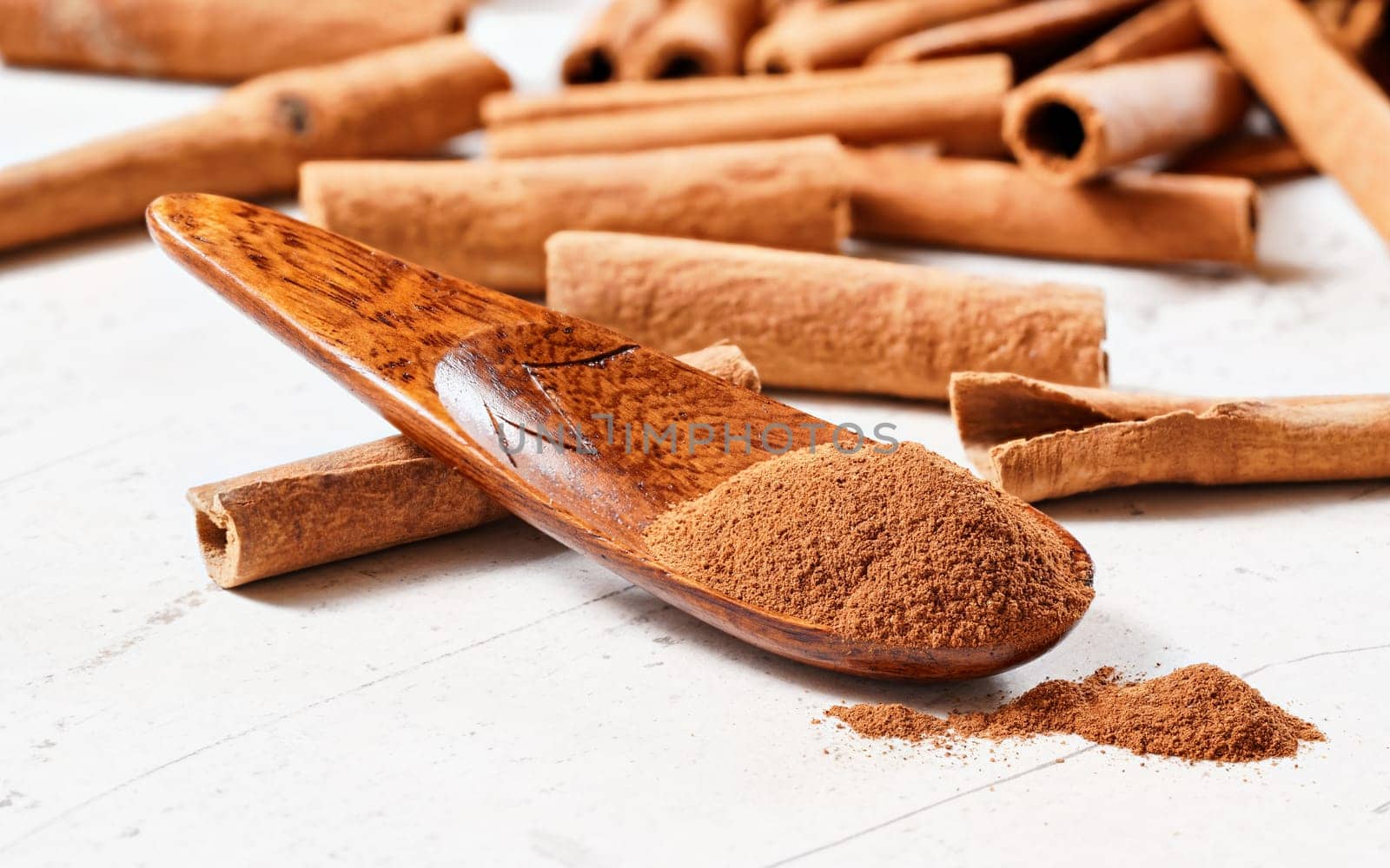 Grounded cinnamon powder in small wooden spice spoon on white board, blurred whole sticks in background, close up macro