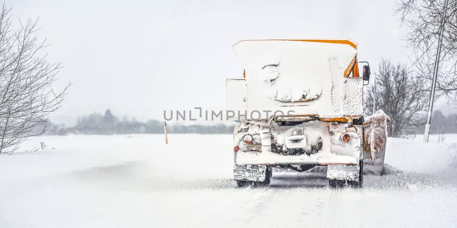 Orange plough truck on snow covered road, gray sky and trees in background, view from back - winter road maintenance by Ivanko