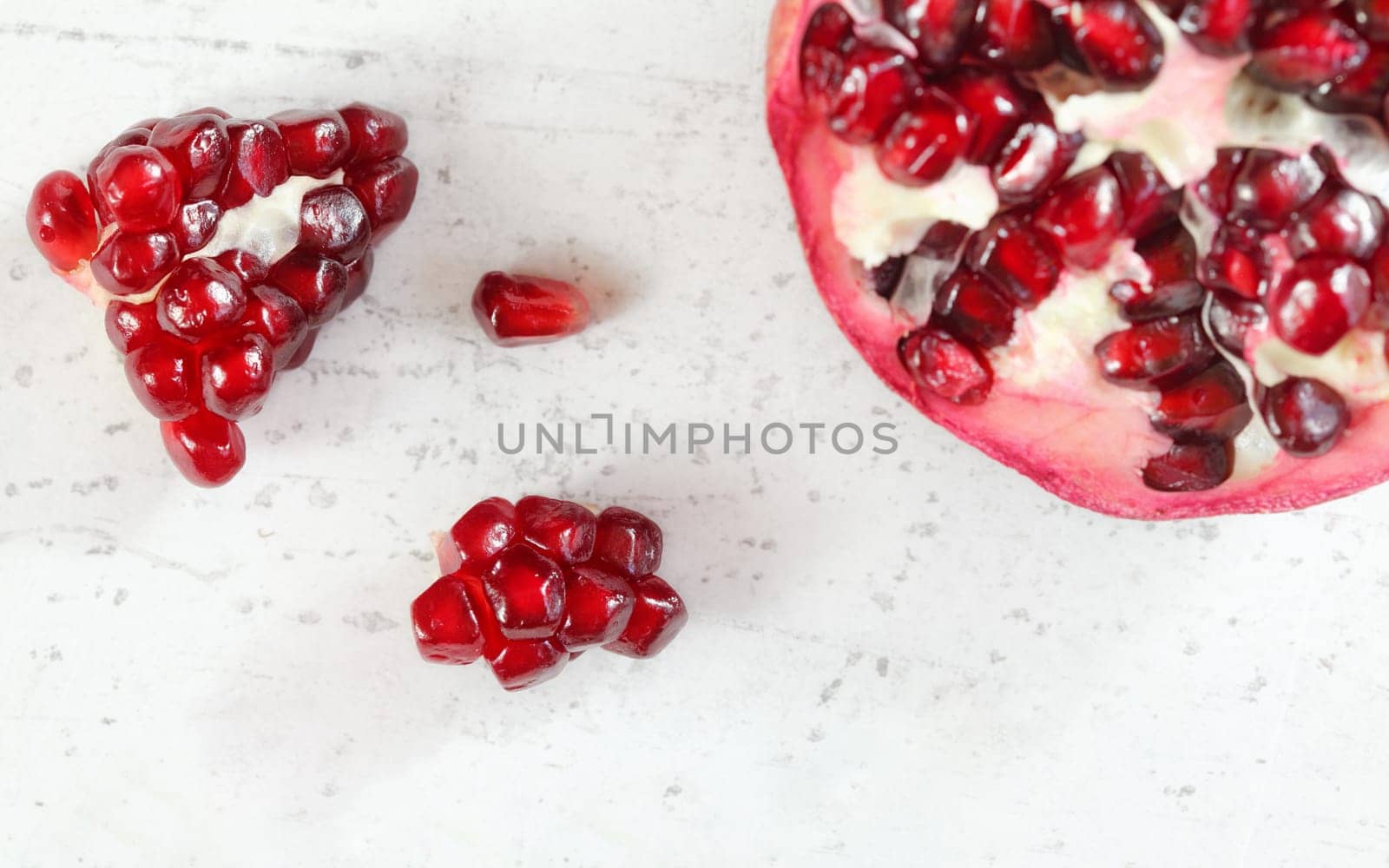 Pomegranate halved, gem like fruits next to it on white stone board, top down view.