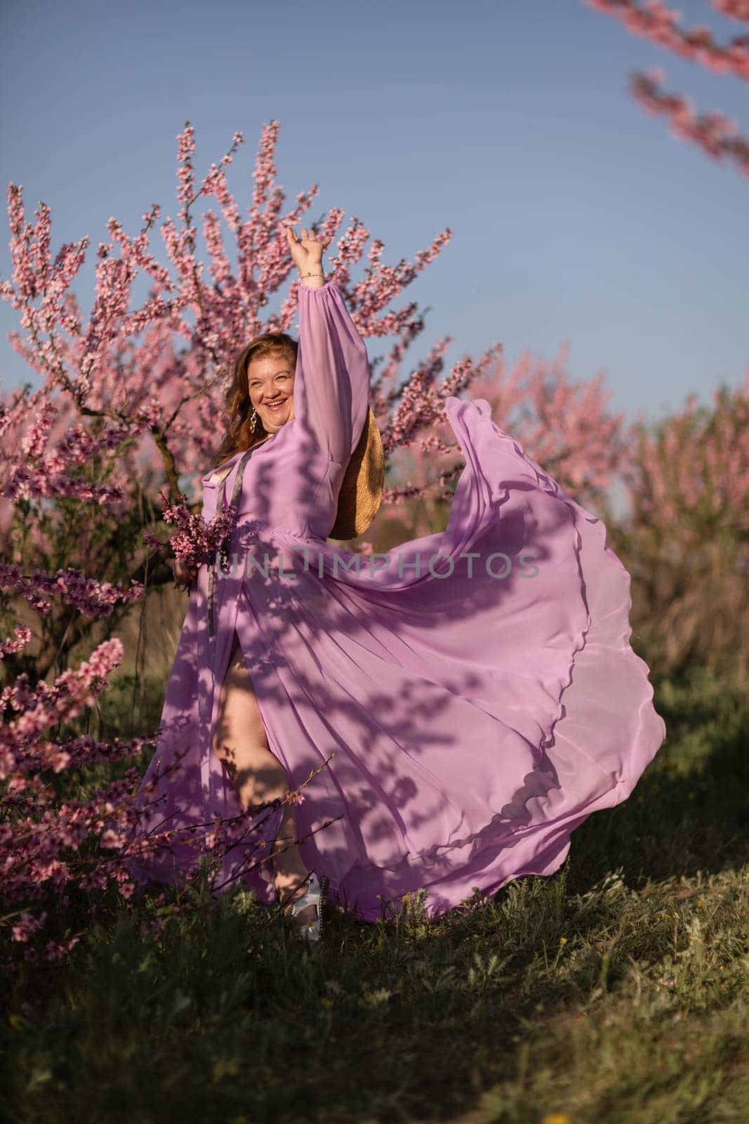 Woman blooming peach orchard. Against the backdrop of a picturesque peach orchard, a woman in a long pink dress and hat enjoys a peaceful walk in the park, surrounded by the beauty of nature