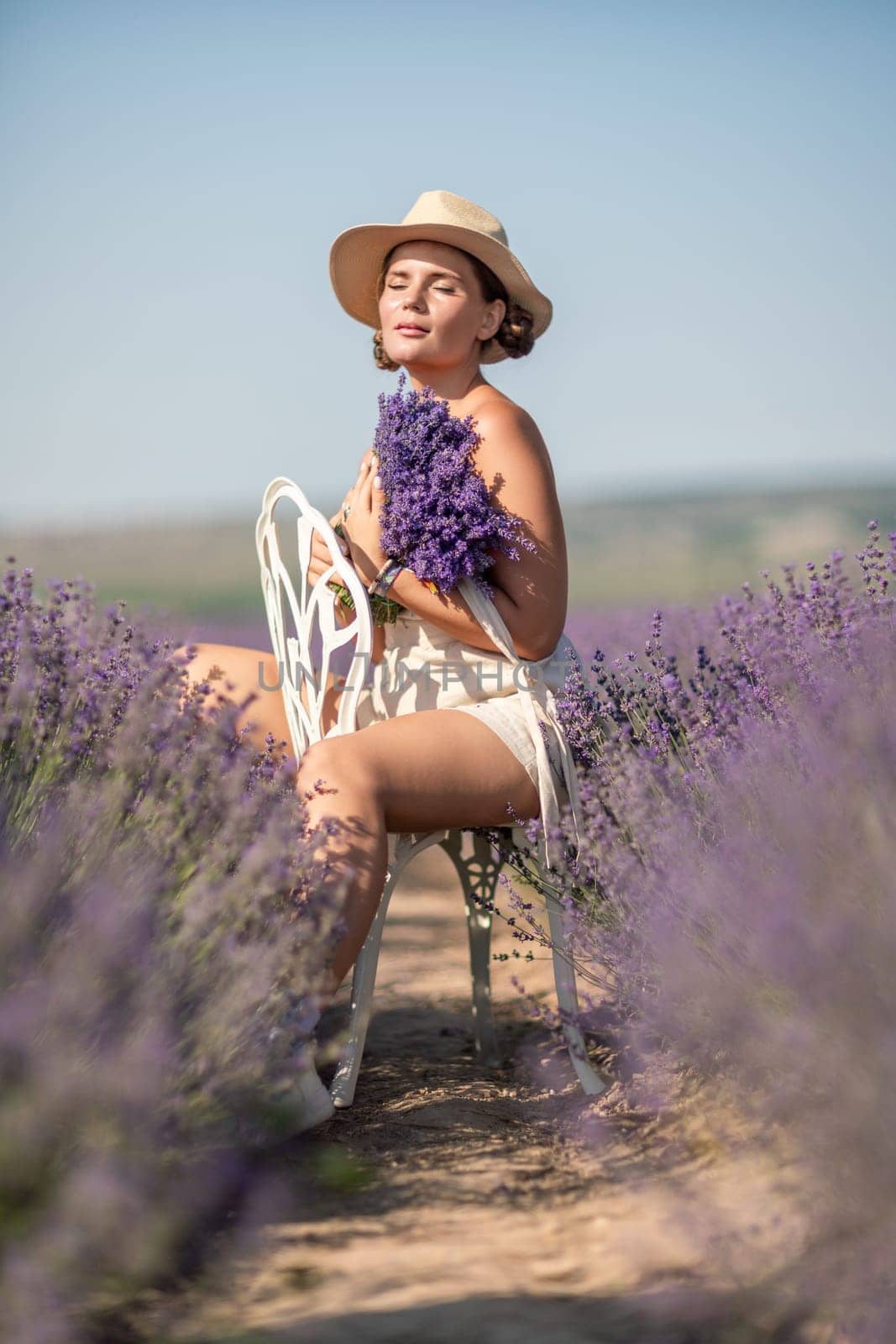 A woman is sitting in a field of lavender flowers and wearing a straw hat. She is smiling and holding a bouquet of flowers. Scene is peaceful and serene, as the woman is surrounded by the beauty of nature.