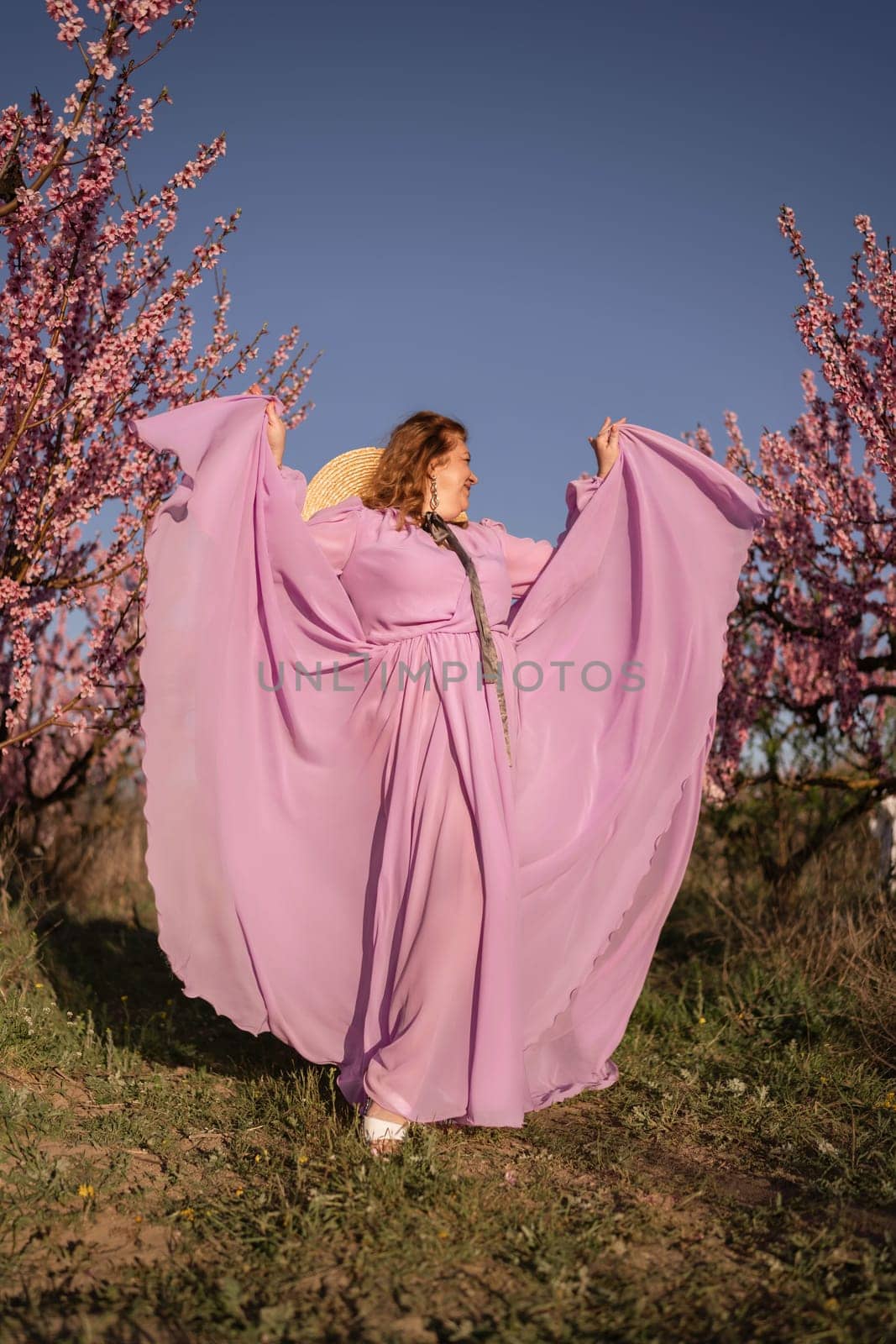 Woman blooming peach orchard. Against the backdrop of a picturesque peach orchard, a woman in a long pink dress and hat enjoys a peaceful walk in the park, surrounded by the beauty of nature