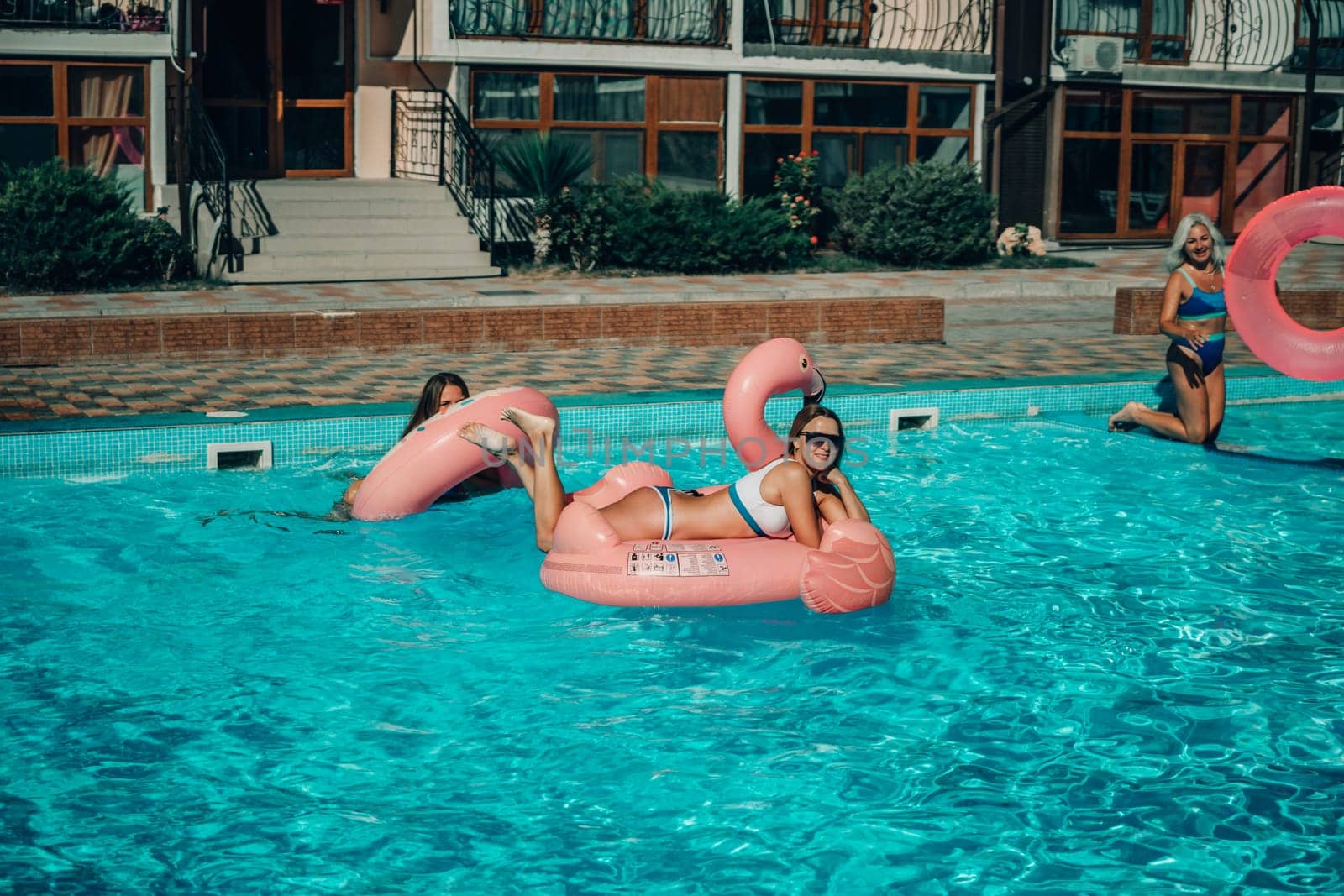 A woman is floating in a pink flamingo float in a pool. The pool is surrounded by a brick wall and has a few other people in the background