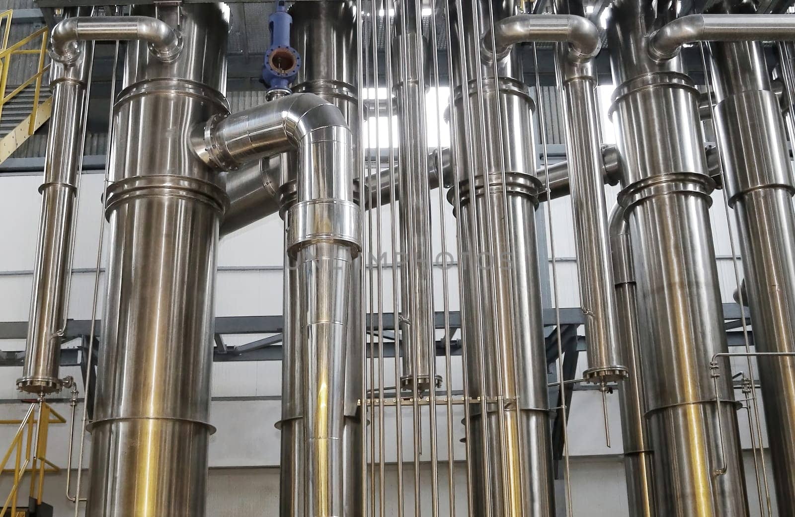 A chemical plant features a complex network of stainless steel pipes and glass vessels, creating an intricate system of interconnected straight and curved components in various sizes and shapes.