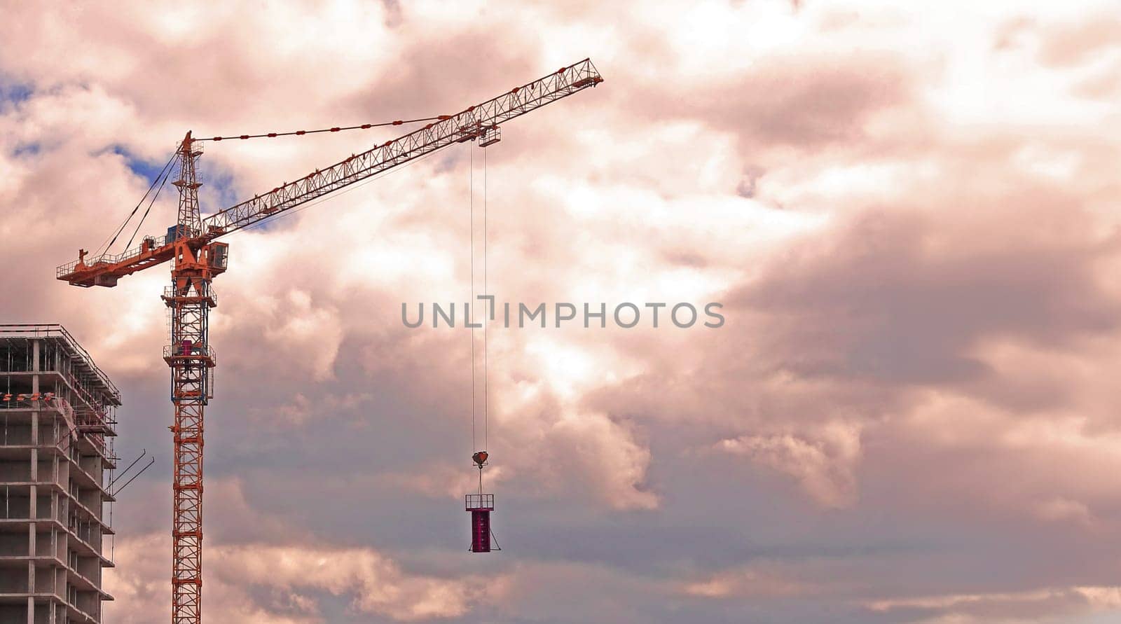 A construction crane stands tall against a backdrop of soft pink and white clouds, with a building under construction visible in the foreground.