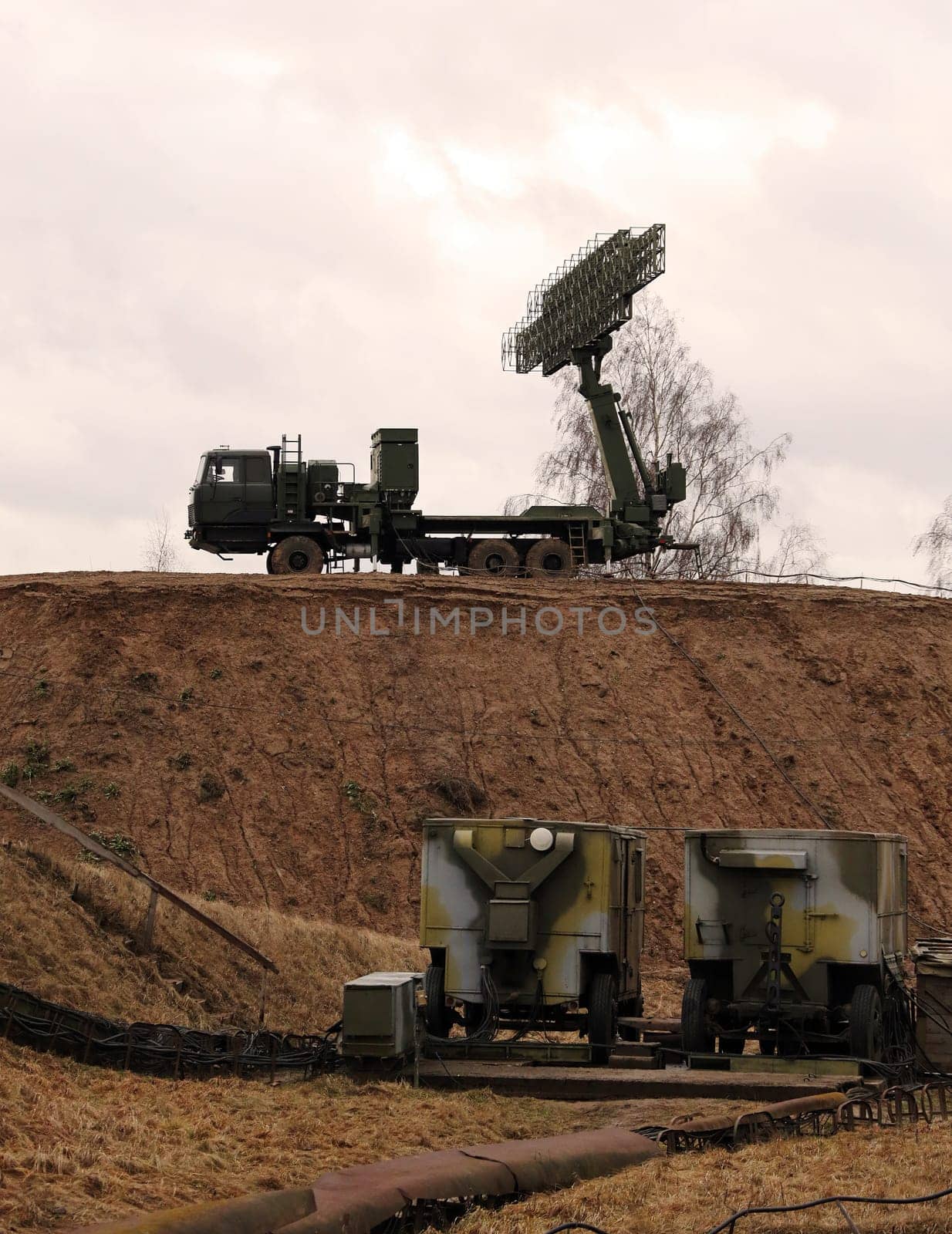 Two large military radar systems, mounted on vehicles, stand on a hill under a cloudy sky.