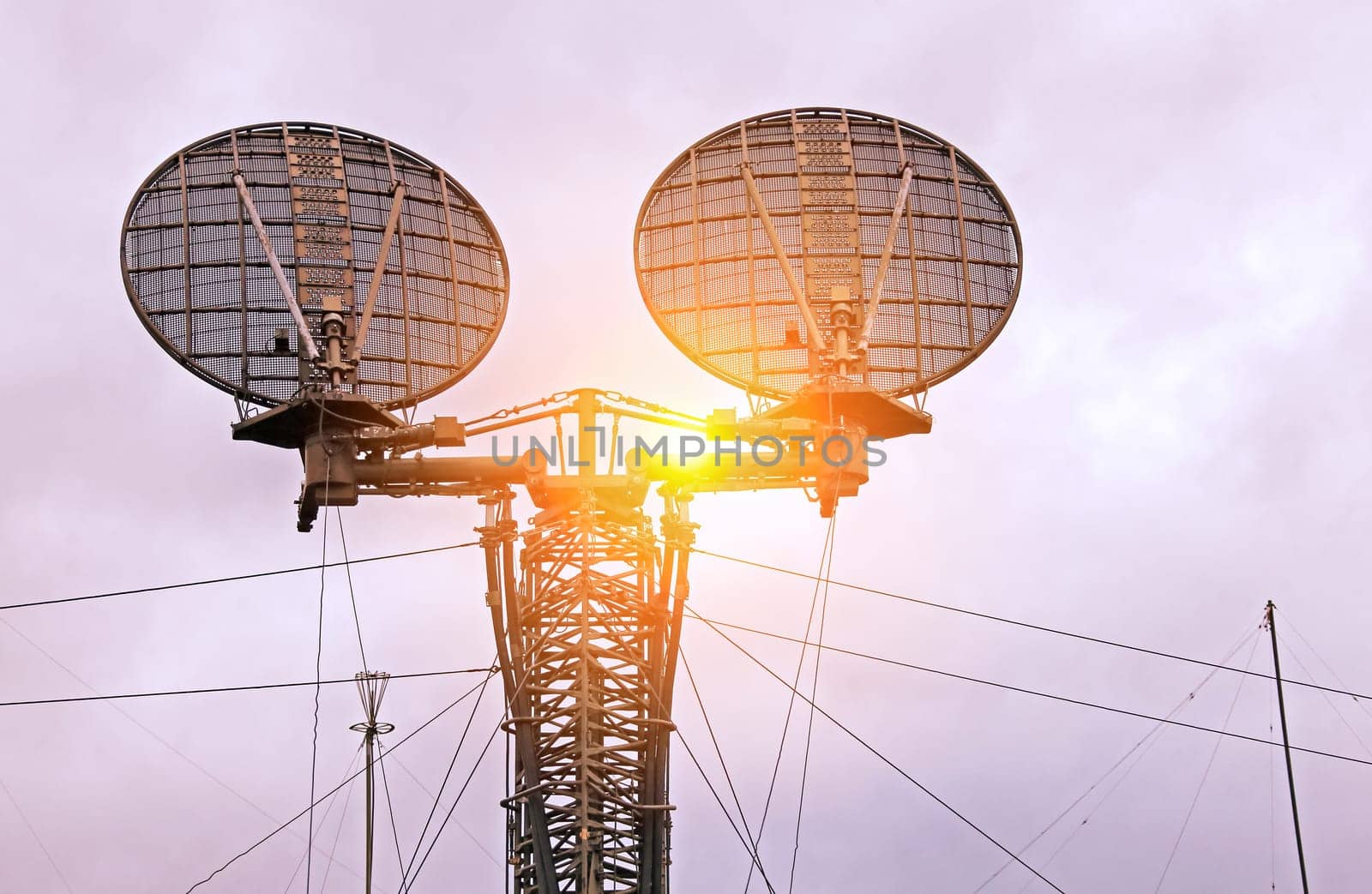 A close-up view of two large radar dishes mounted on a tall tower, with a cloudy sky in the background.