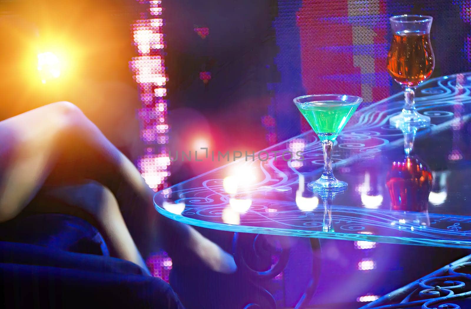Close-up shot in dimly lit nightclub: table with green and orange cocktails under neon lights, persons legs in foreground.
