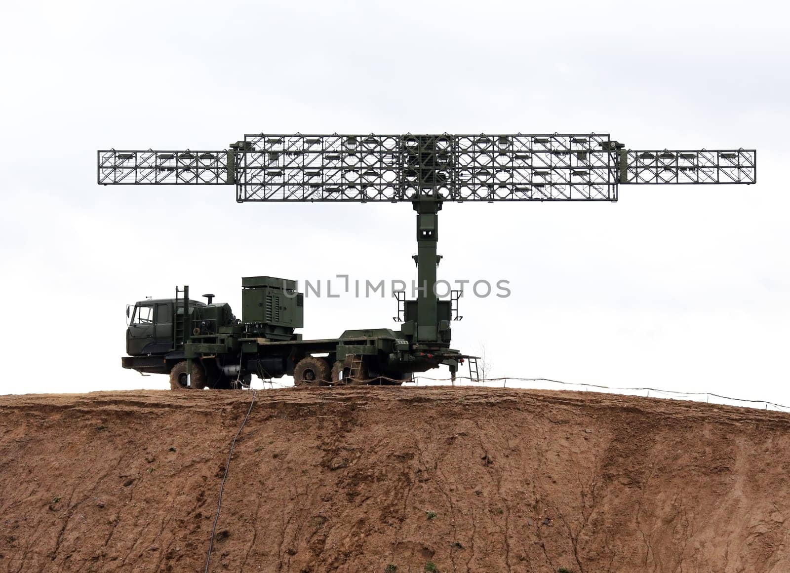A large, military radar system is mounted on a truck and positioned on a dirt hilltop. The radar dish is extended and pointed upwards, likely scanning for potential threats in the sky.