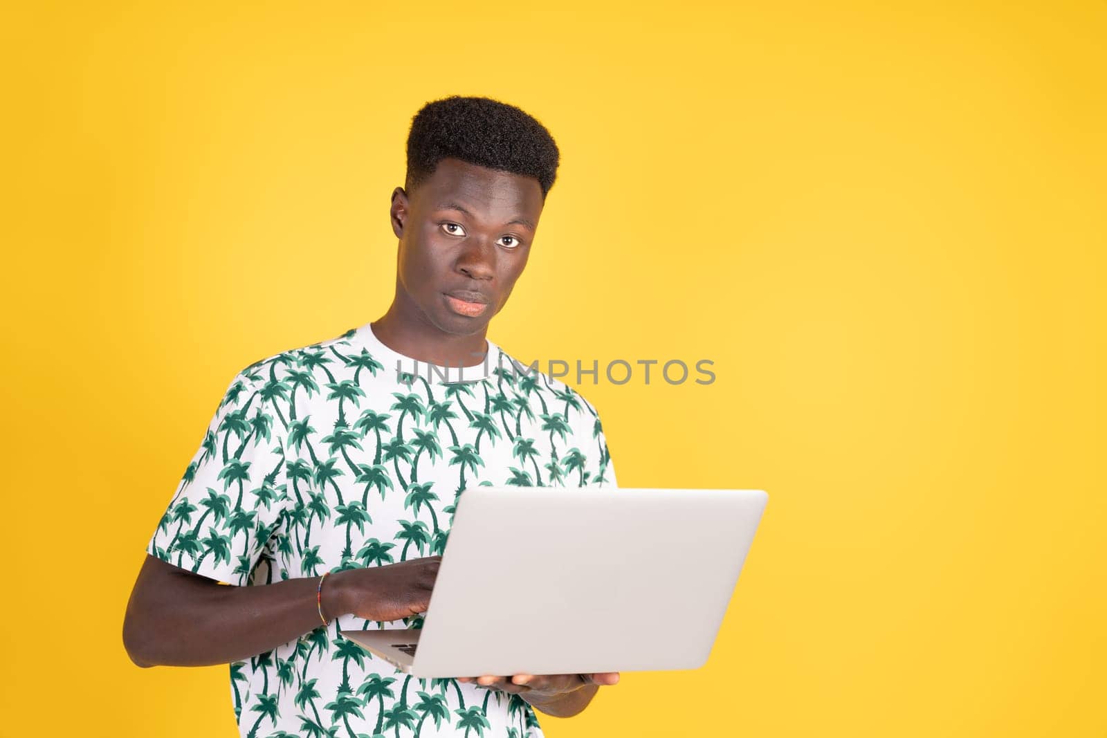 Young man with serious look of African ethnicity wearing summer t-shirt with palm trees working on a laptop in a studio portrait with the yellow background. People lifestyle concept.