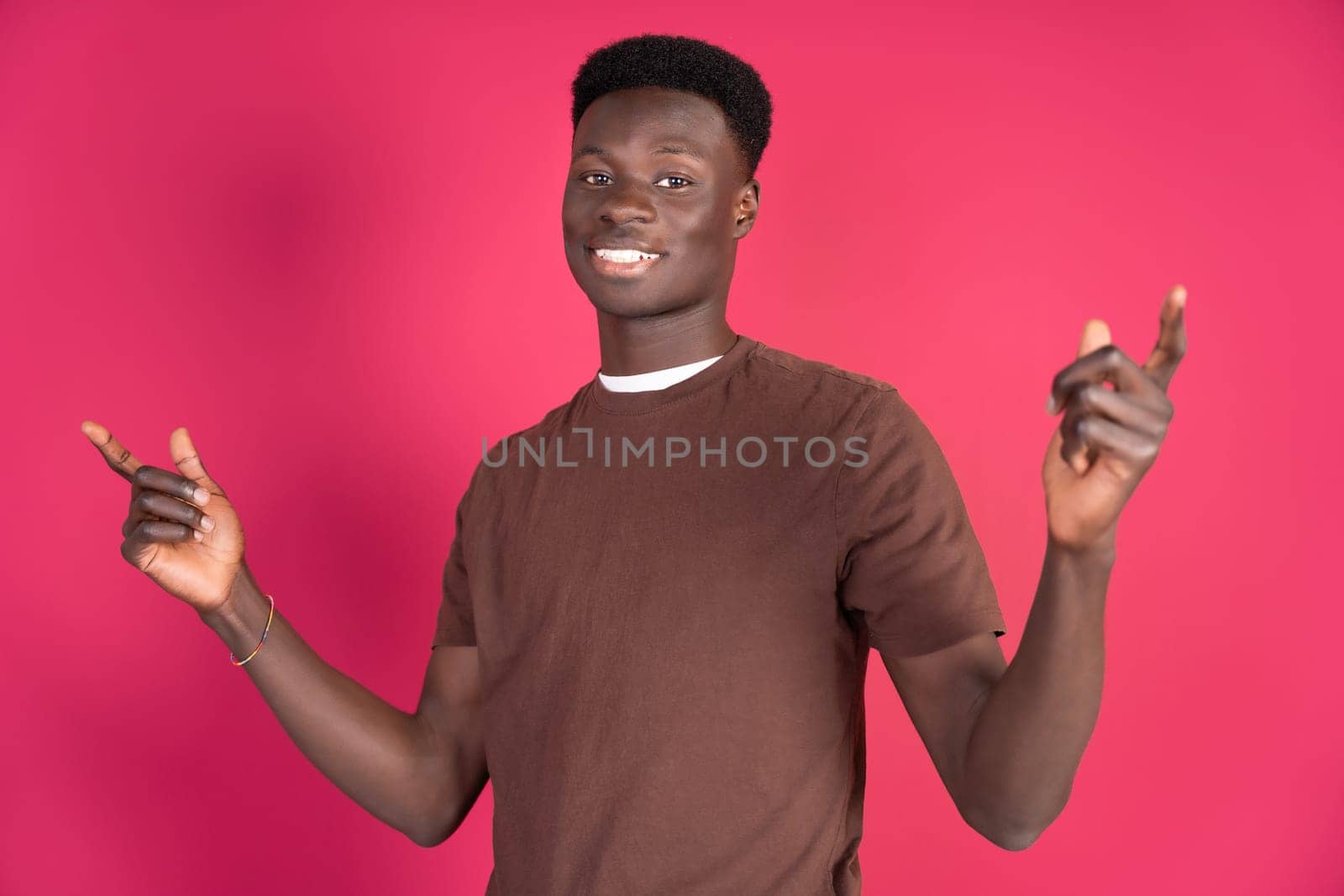 A cheerful young African man stands against a vibrant pink backdrop, pointing upwards with both hands. His bright smile and casual brown T-shirt suggest a lively and engaging atmosphere. The man appears to be in the midst of a conversation or presentation, using hand gestures to direct attention above.