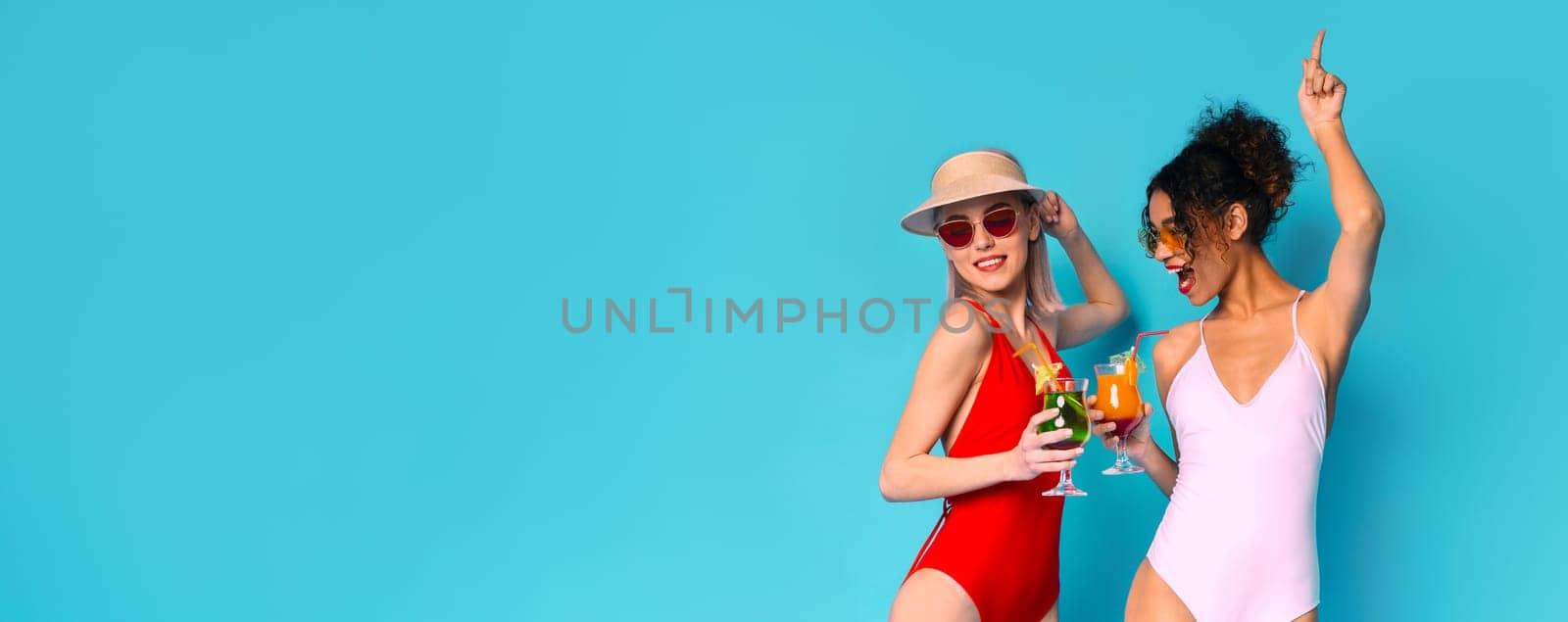 Two women in swimsuits are celebrating with drinks in their hands. They are standing in front of a bright blue background, panorama with copy space
