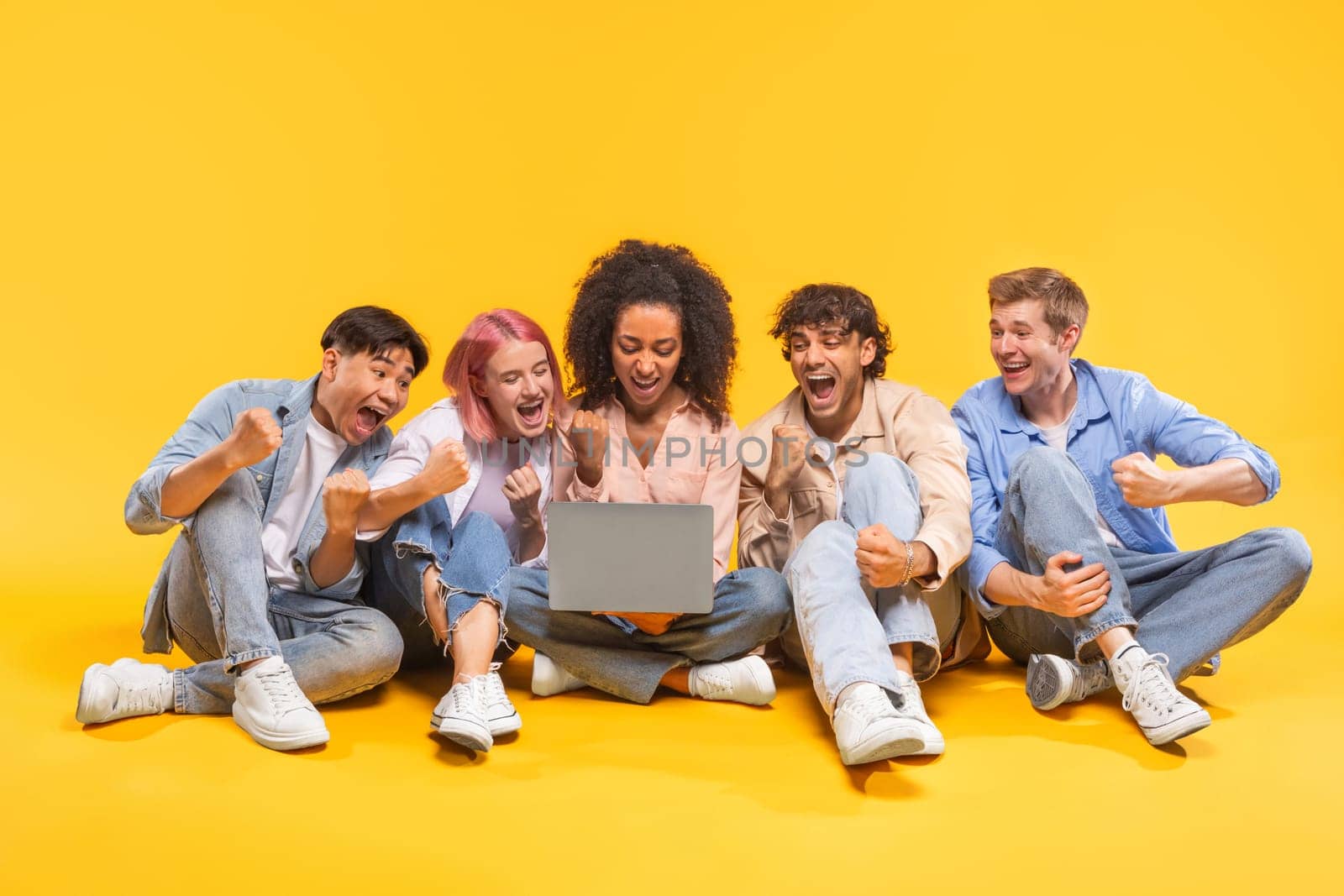 A group of five diverse friends are sitting on a yellow background, looking at a laptop and celebrating. They are all smiling and have their hands in the air.