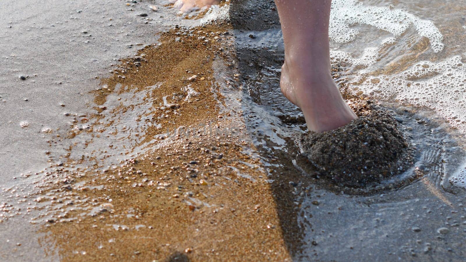 Female feet barefoot in waves of foamy water on a sandy beach on a summer day, top view of female feet, woman on the beach getting her feet wet in warm sea water, carefree vacation by the sea,
