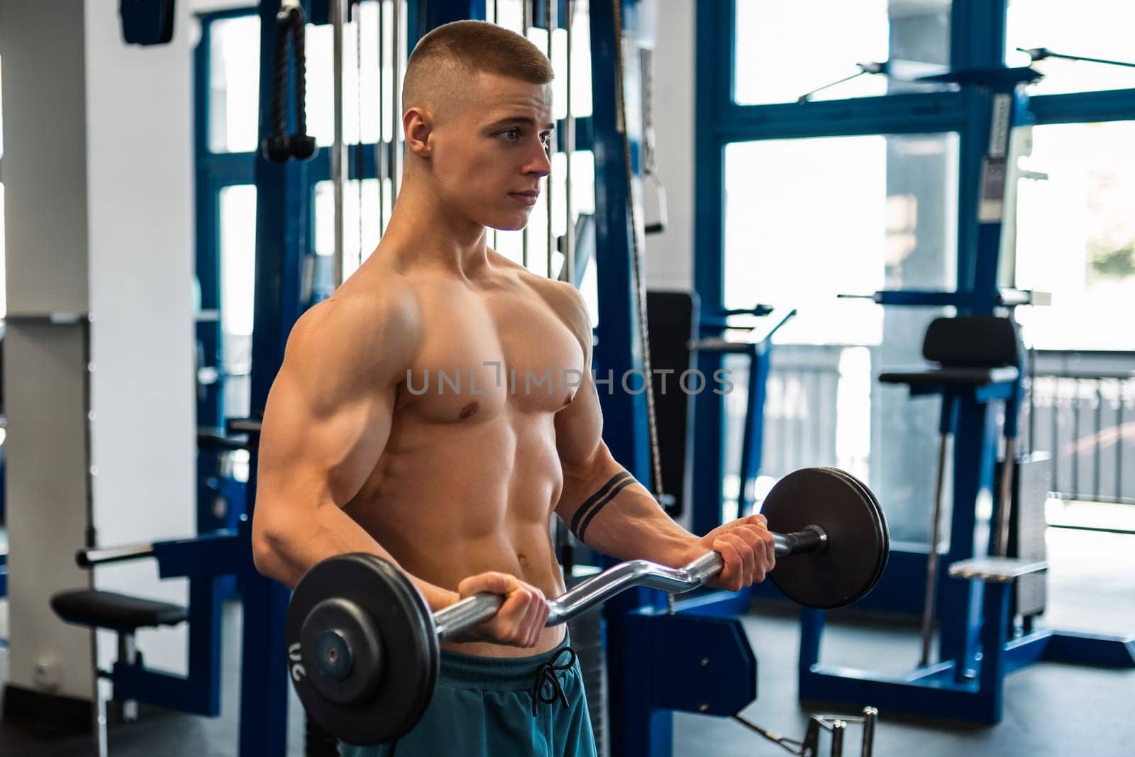 A shirtless bodybuilder trains his biceps using a barbell in a gym