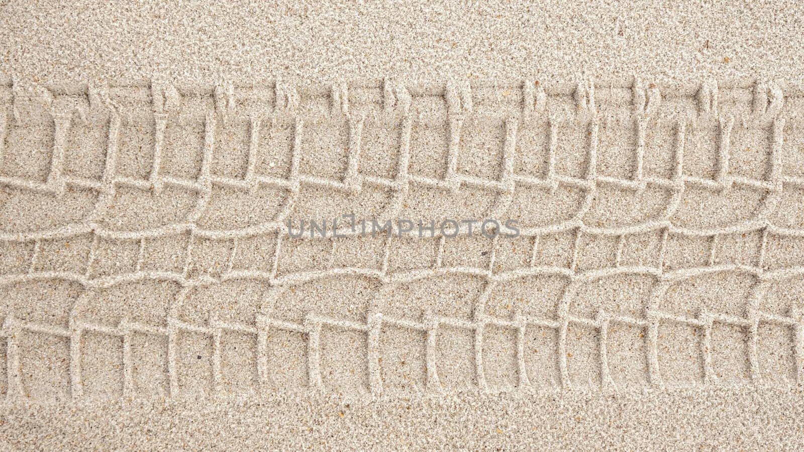 A detailed view of a wide tire track in light brown sand, likely from a heavy vehicle. The dark brown track, approximately 12 inches wide, shows human impact on natural landscapes.