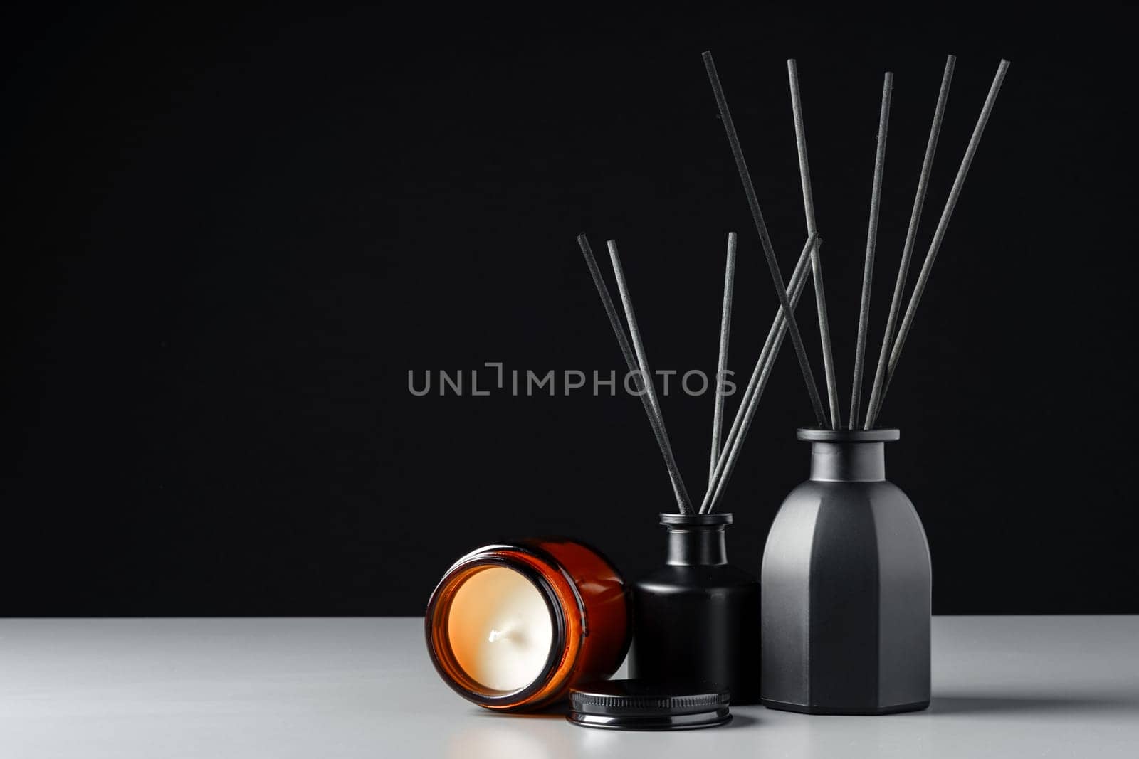 A black reed diffuser bottle with black reeds is placed on a grey surface with a candle next to it. The background is a solid black.