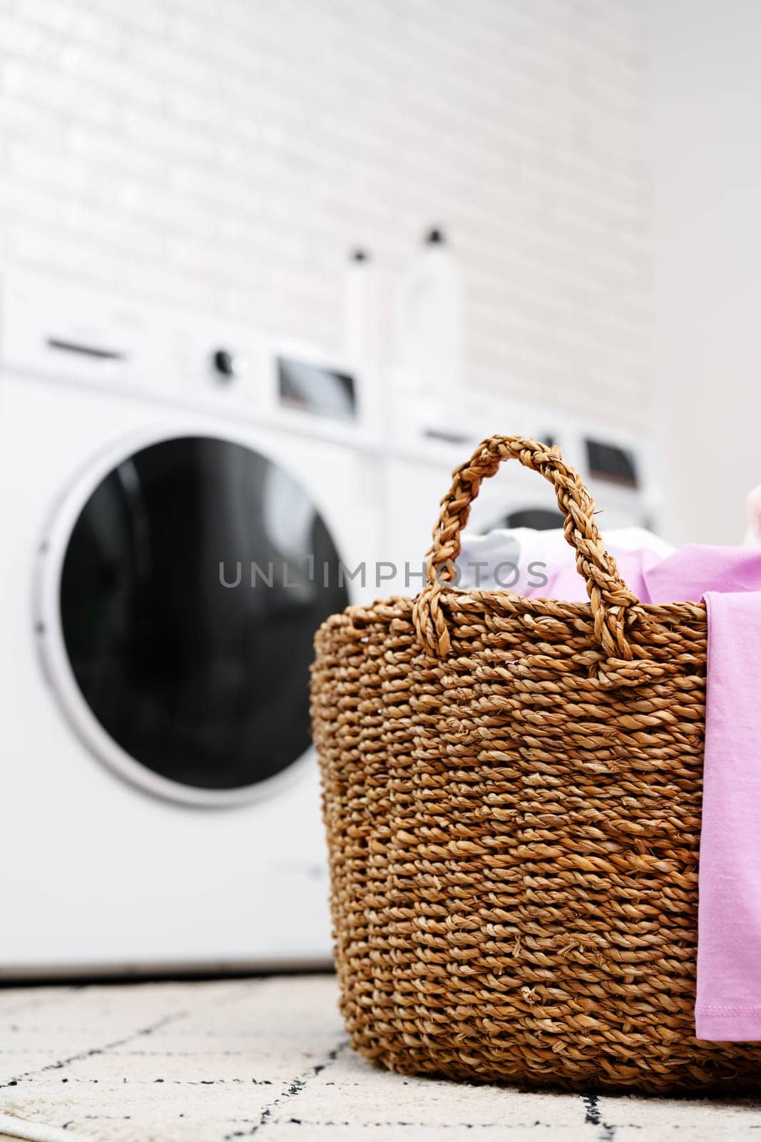 A wicker laundry basket filled with clean, folded clothes sits on a rug in front of two modern washing machines in a bright, clean laundry room.