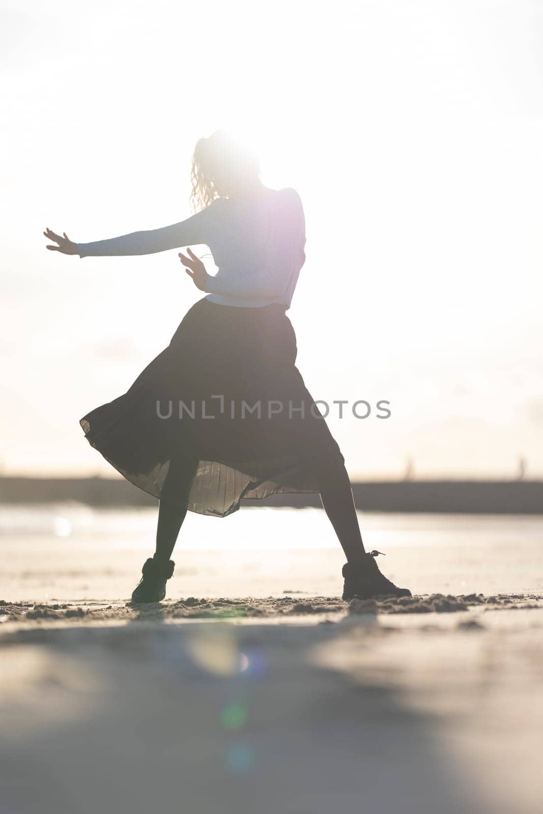 A woman is dancing on the beach in a skirt. The image has a light and playful mood, as the woman is dancing in the sun and the beach setting is bright and cheerful