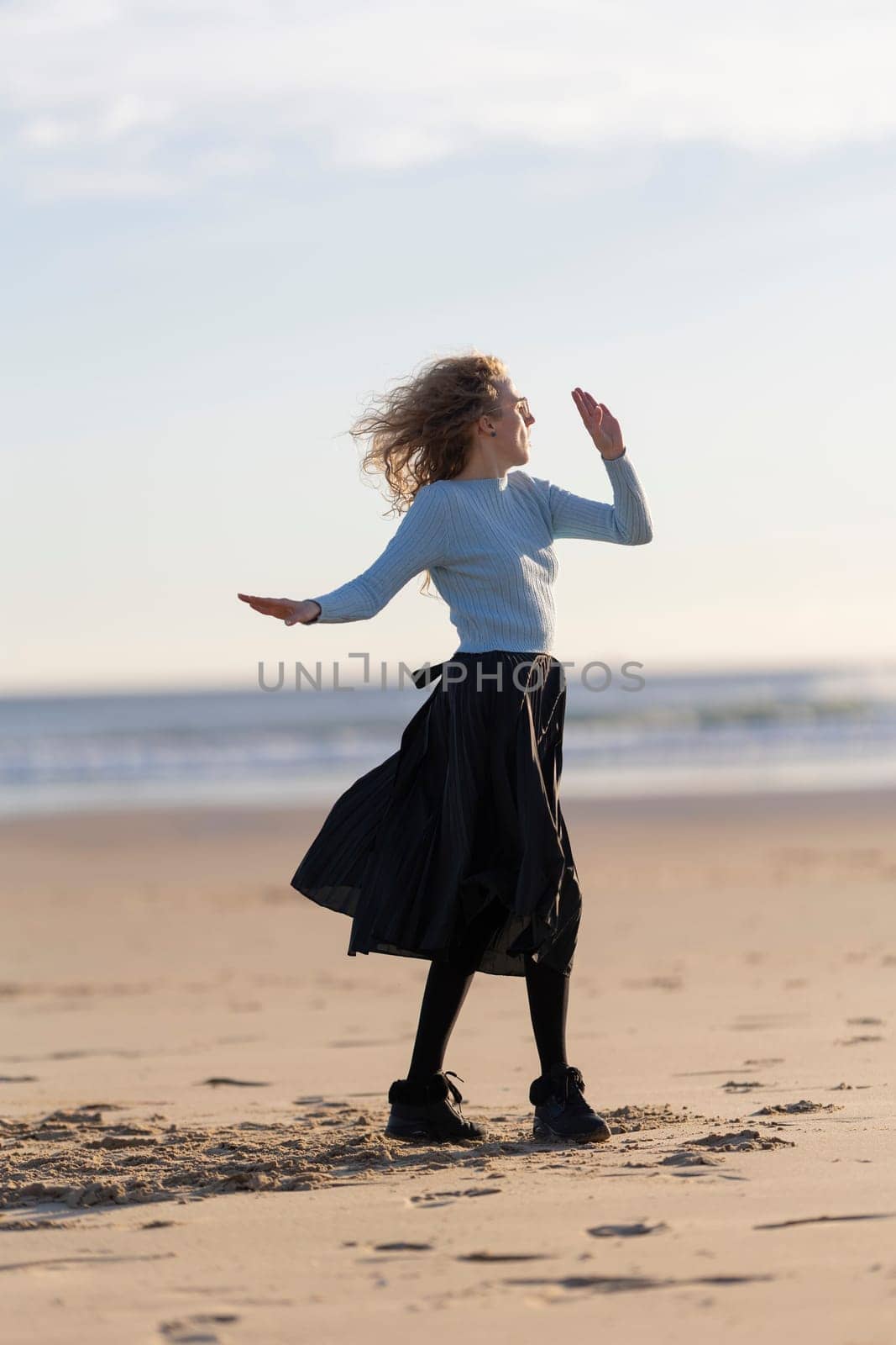 A woman is standing on the beach, wearing a blue shirt and black skirt. She is dancing and she is enjoying herself. The beach setting and the woman's carefree attitude create a sense of relaxation