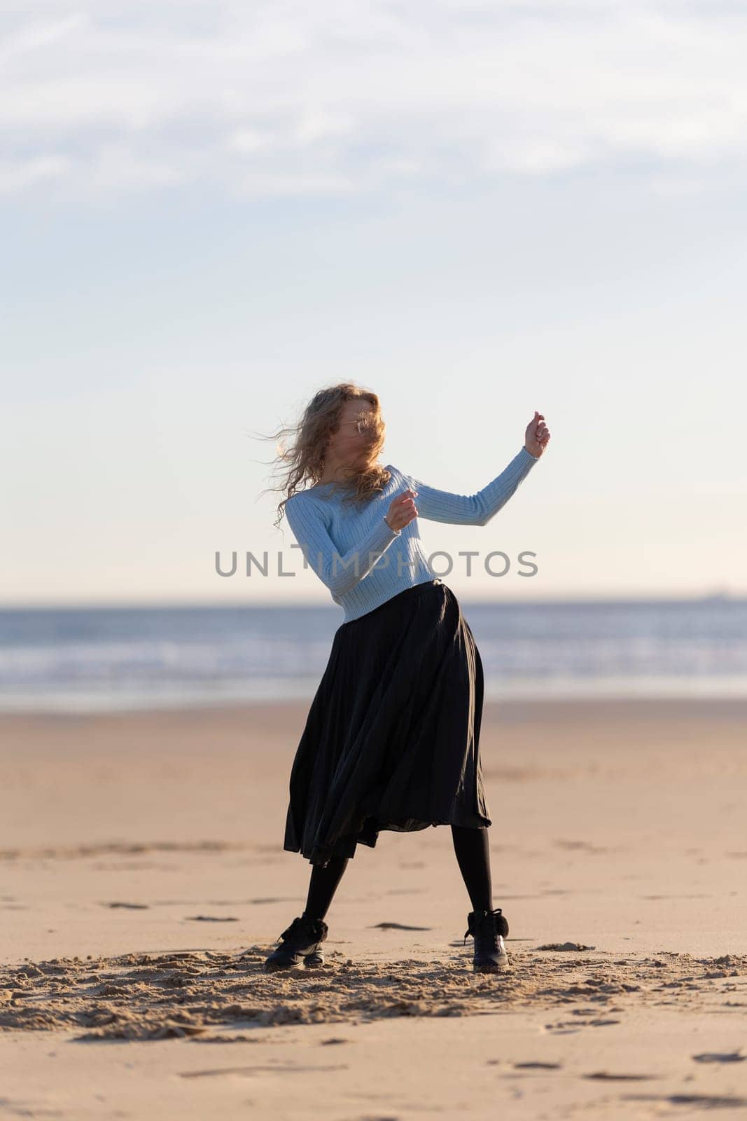 A woman is dancing on the beach in a black skirt. She is wearing a blue shirt and black boots