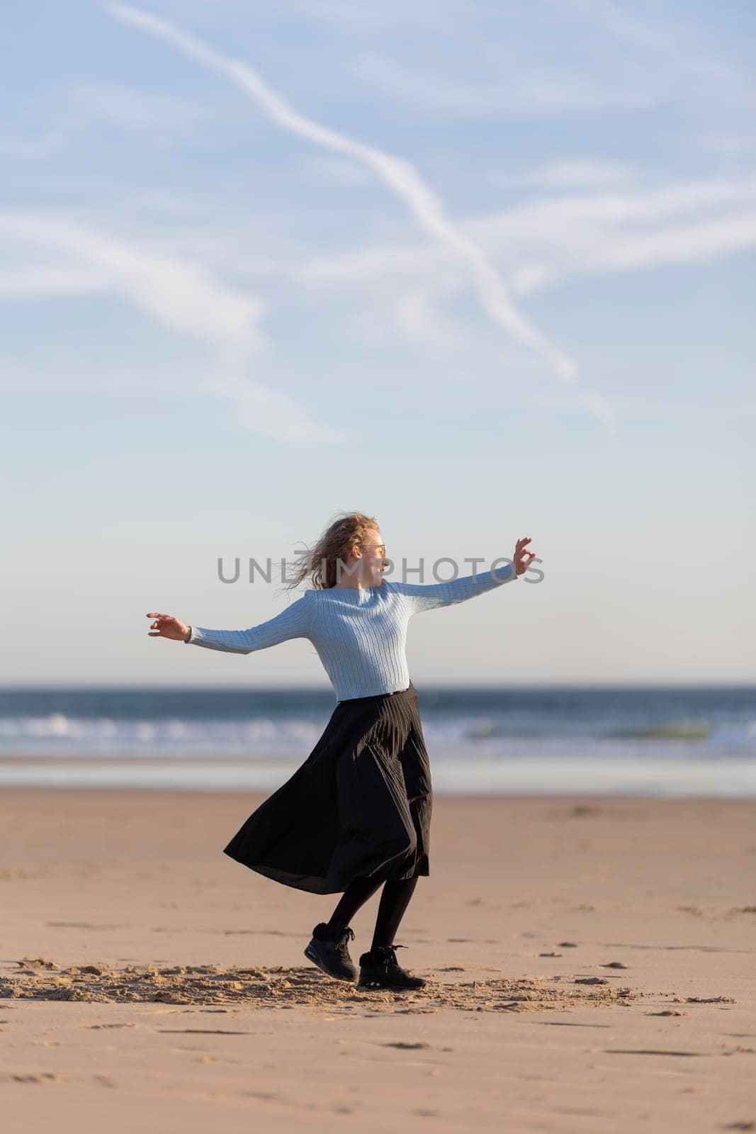 A woman is standing on the beach, wearing a blue shirt and black skirt. She is smiling and she is enjoying herself. The beach is a beautiful and peaceful setting, with the ocean in the background
