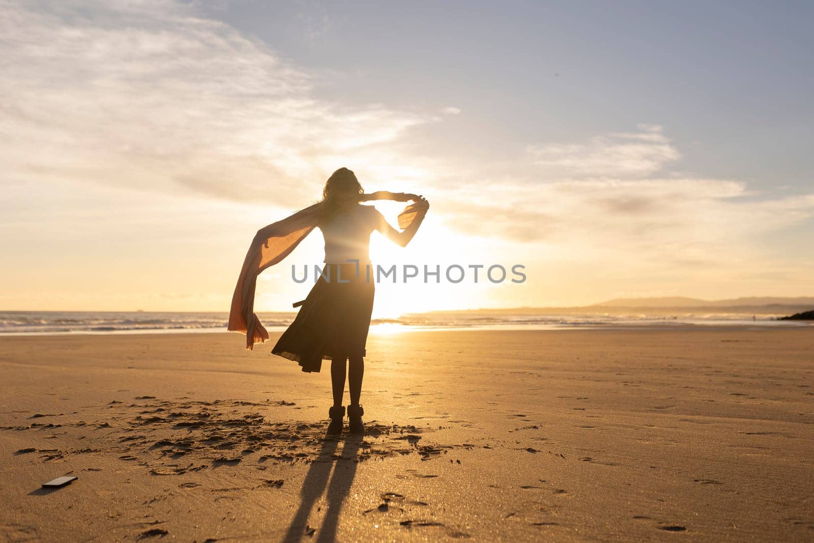A woman stands on a beach with a scarf around her neck. The sky is a beautiful orange color, and the sun is setting. The woman is enjoying the moment and the beautiful scenery